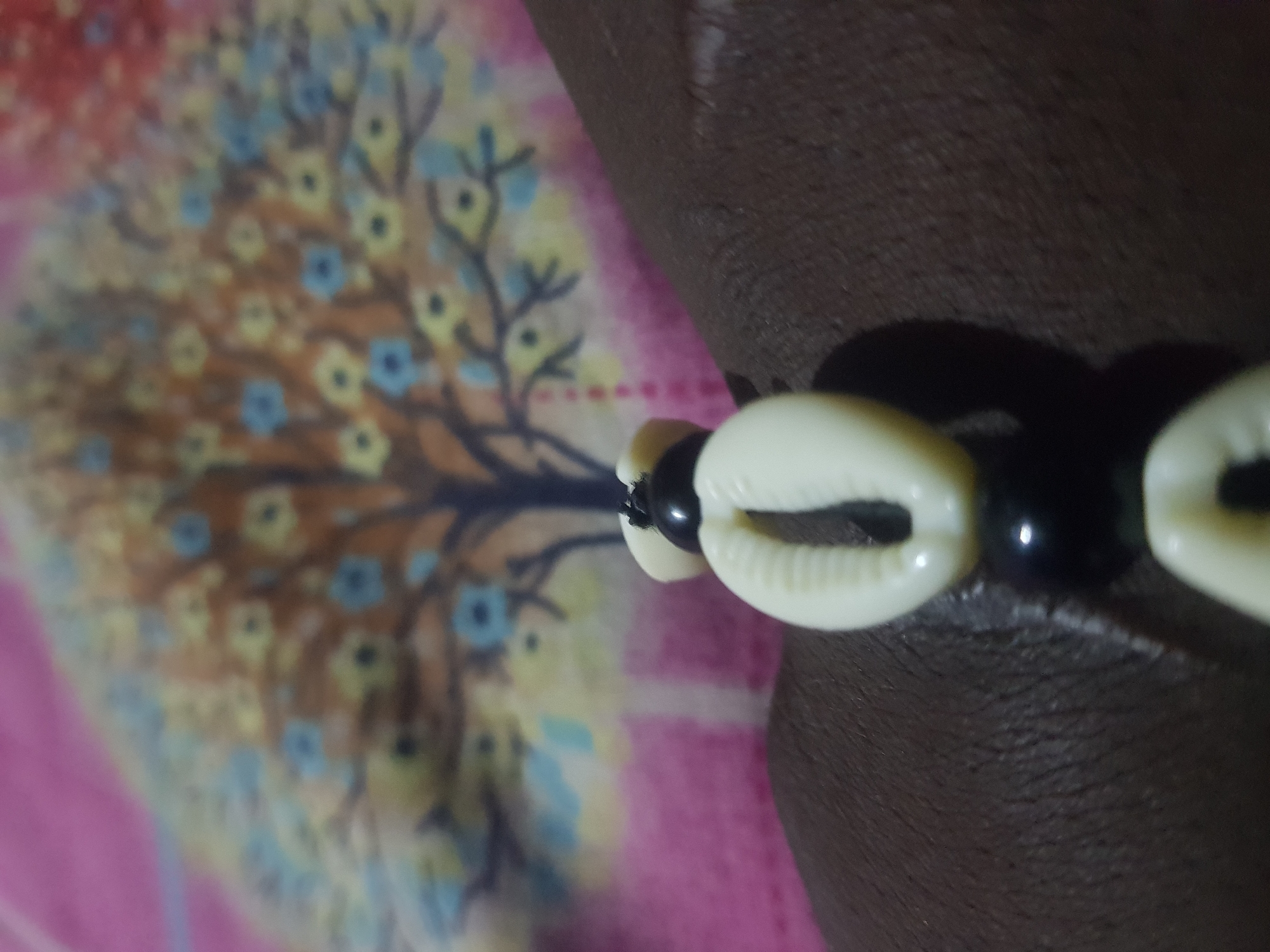 A close-up shot shows a bracelet on a person's wrist. The bracelet features black beads and cowrie shells. In the background, a blurred image depicts a colorful tapestry or fabric with a tree motif. The image is taken from an unknown location in an unknown city.