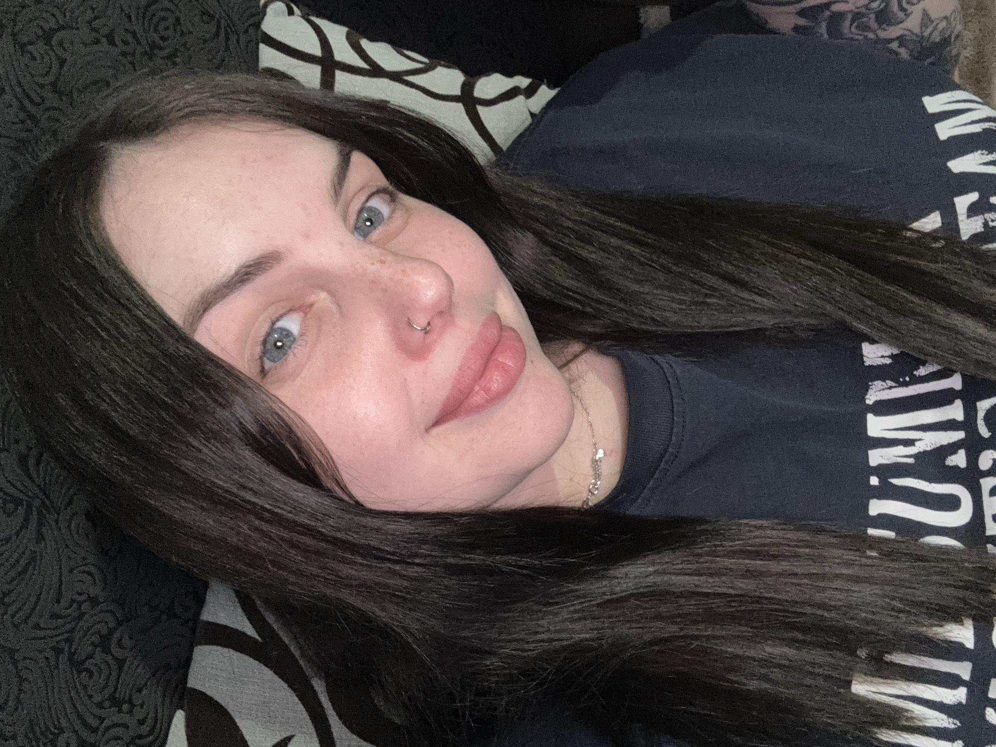 The image shows a woman with long, dark hair and fair skin. She has light-colored eyes and a nose piercing. She is lying down, with her head resting on a patterned surface that appears to be a cushion or pillow. She is wearing a dark-colored t-shirt with white text. The background is blurry and does not offer any clear indication of a location in Chornomorskoe, Ukraine, or any other specific location. There is no visible interaction or action taking place. The environment appears to be indoors.