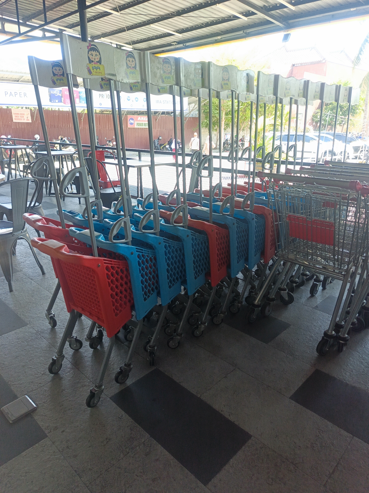 In Gorontalo, Indonesia, a row of blue and red shopping carts are lined up beneath an open-air shelter, possibly at a market or supermarket. The carts are mostly empty, with some showing signs of use. Behind the carts, tables and chairs are visible, along with parked motorcycles and several cars. A partially obscured sign with a cartoon character and text in Indonesian hangs above the carts. The ground is paved with tiles, and a silver object resembling a phone lies on the floor in the foreground. The overall scene suggests a commercial area on a sunny day.