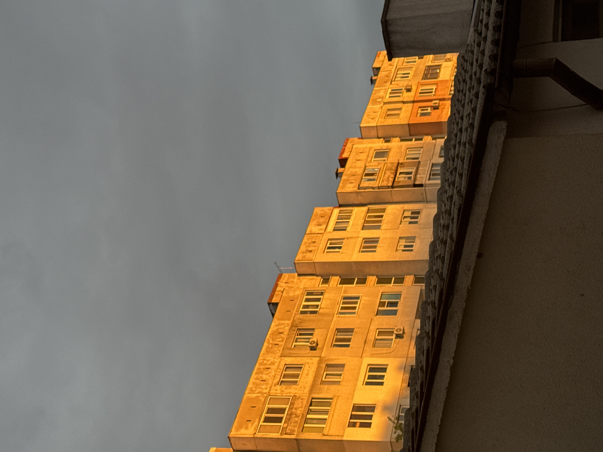 The image shows a high-rise building in Unknown, Unknown. The building has a stepped facade, with sections that appear to stack upon one another. The exterior is lit with a golden light, suggesting it may be sunrise or sunset. The sky is overcast and gray. The perspective is looking up at the building from street level, with a portion of another building visible in the foreground on the right. There are numerous windows visible in the facade. There are air conditioning units installed in some of the windows.