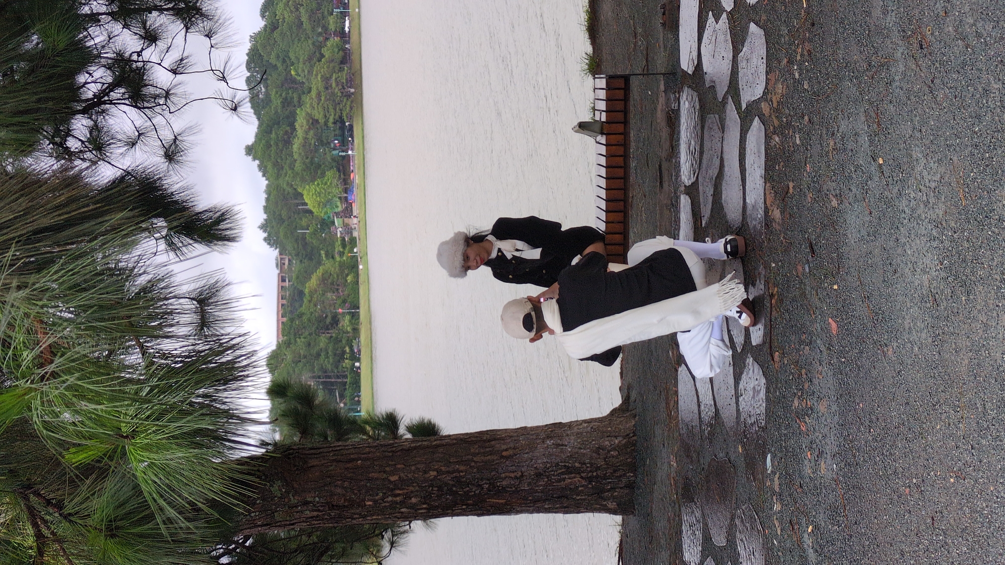 In Da Lat, Vietnam, by a lake with a tree in the foreground and a bench by the water, a person kneels on the ground, holding up a smartphone to take a picture of another person. The person kneeling is wearing a black top, white scarf, white pants, a white cap, and white socks with brown and black shoes. The person on the bench is wearing a black jacket, white scarf, white hat, and black shoes. They are looking at the person taking the photo. The lake is calm and reflects the cloudy sky. The surrounding area is covered with grey gravel and large, irregular stone pavers. In the background, a lush green forest extends to the horizon, with a large building visible amongst the trees.