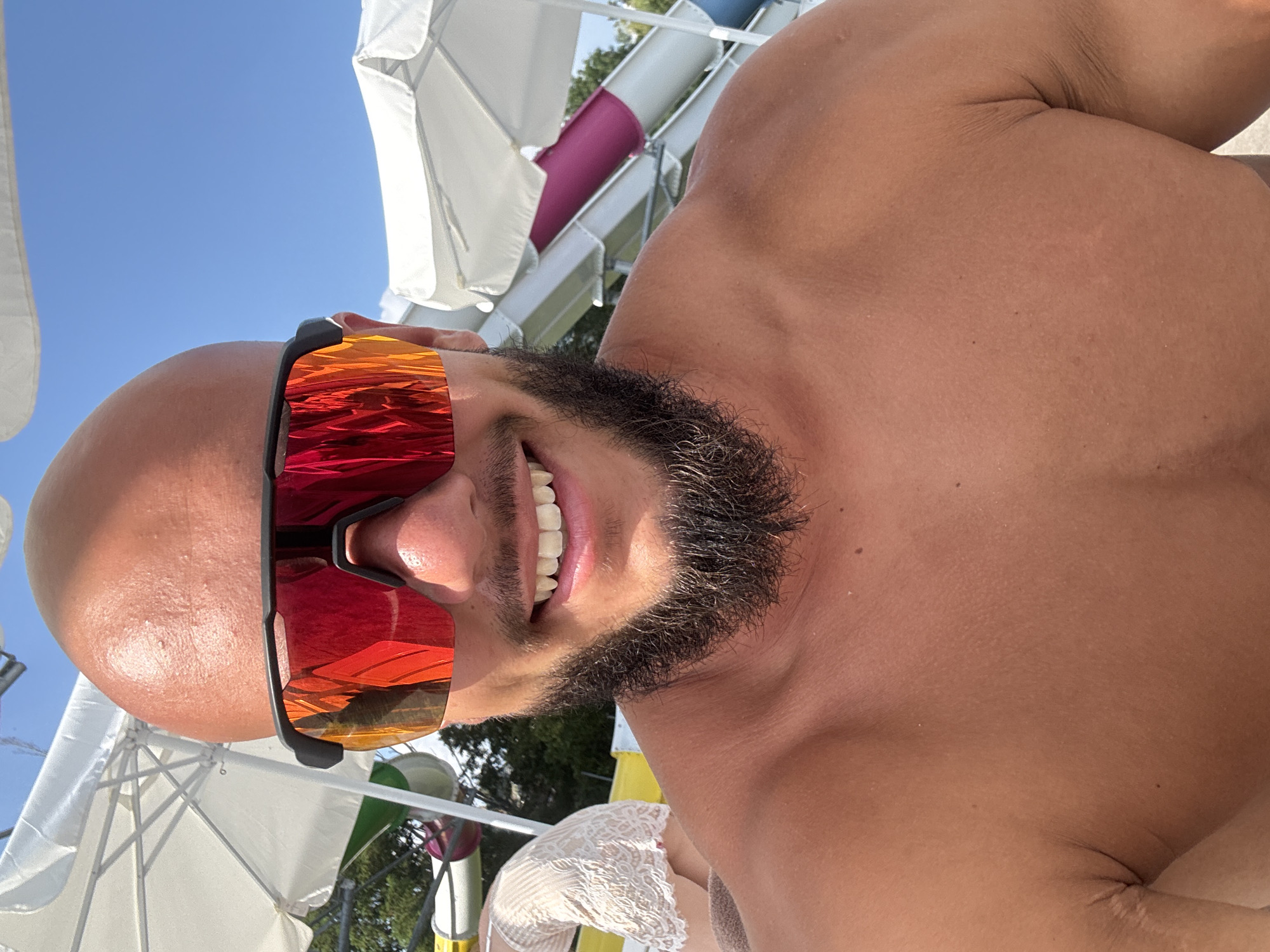 In Ivanyane, Bulgaria, a man with a shaved head and a beard smiles broadly, wearing mirrored sunglasses. He is shirtless, exposing his tanned chest and shoulders. Behind him, a water slide complex is visible with white umbrellas providing shade. A person in a white lace top is partially visible on the left, seated near a yellow water slide. The sky above is a clear blue, indicating a sunny day.