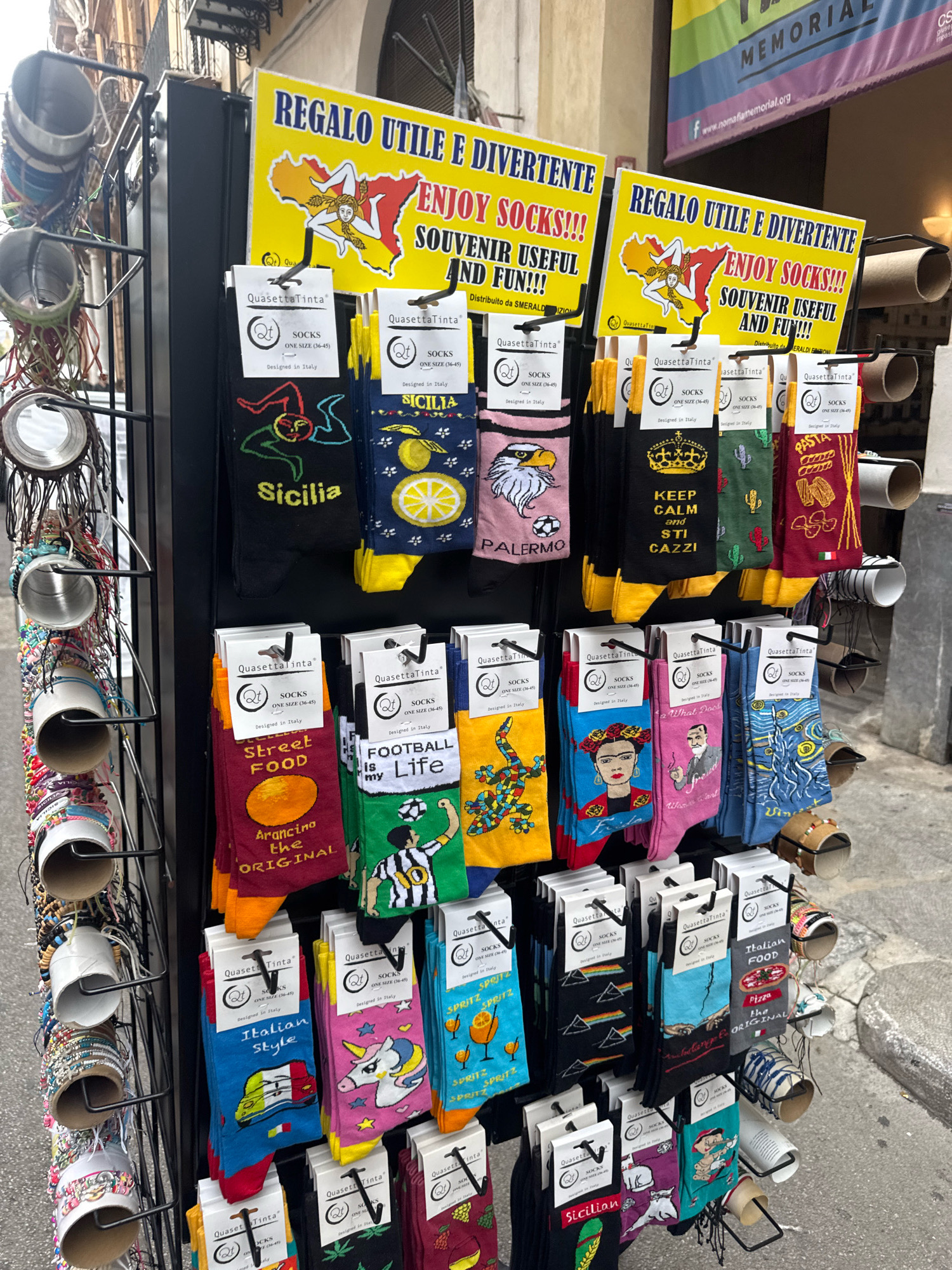 This image shows a display of novelty socks for sale outside a shop in Palermo, Italy. The socks are packaged in clear plastic sleeves with a cardboard header that reads "Quasatta Tinta," "SOCKS," and "ONE SIZE (36-45) - Designed in Italy." The sock designs feature various themes, including local motifs like the Trinacria symbol (a woman's head with three serpent legs), Sicilian landmarks, and popular Italian foods like pasta and pizza. Other designs include abstract patterns, cultural references like Frida Kahlo and Van Gogh, and humorous slogans.
The display stand is a black metal rack with multiple tiers holding the socks. Above the socks, two yellow signs with red and black text read "REGALO UTILE E DIVERTENTE ENJOY SOCKS!!! SOUVENIR USEFUL AND FUN!!!". One of these signs prominently features the Trinacria symbol.
To the left of the sock display, there is a rack of bracelets and beaded items. In the background, a banner with purple, green, and white stripes is partially visible, with text that appears to be related to a memorial event. The ground is paved with grey stones, suggesting an outdoor market or street setting. The overall impression is a vibrant, tourist-oriented retail display in a Mediterranean city.