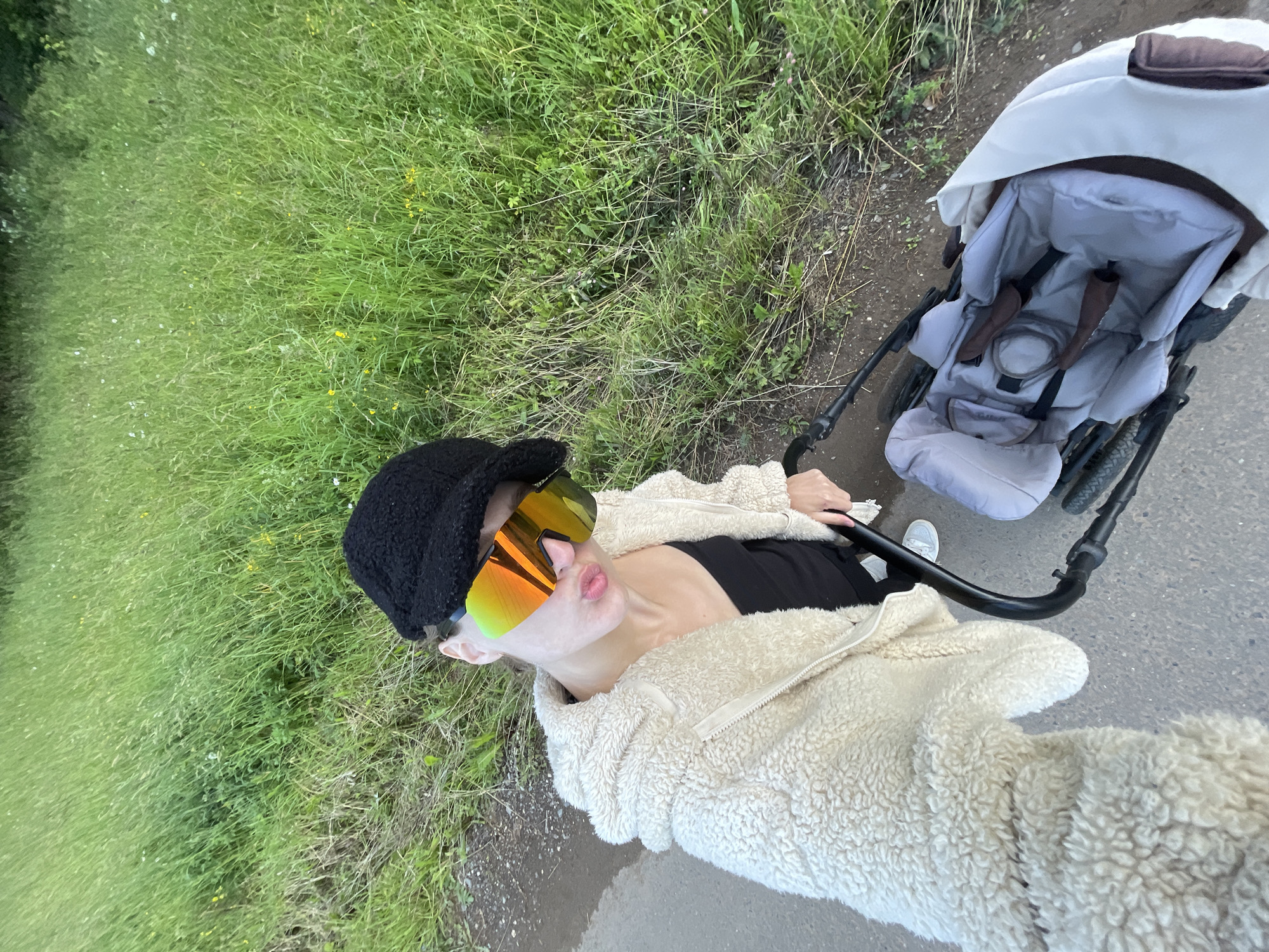 The image shows a woman wearing a black cap, oversized reflective sunglasses, and a cream-colored fuzzy jacket. She is holding a stroller with a light gray interior and brown accents. The woman is standing on a paved path next to a grassy embankment. The background consists of tall green grass with some yellow wildflowers. The scene is set in an outdoor environment, possibly a park or a rural area, near the village of Podshivalovo, Russia.