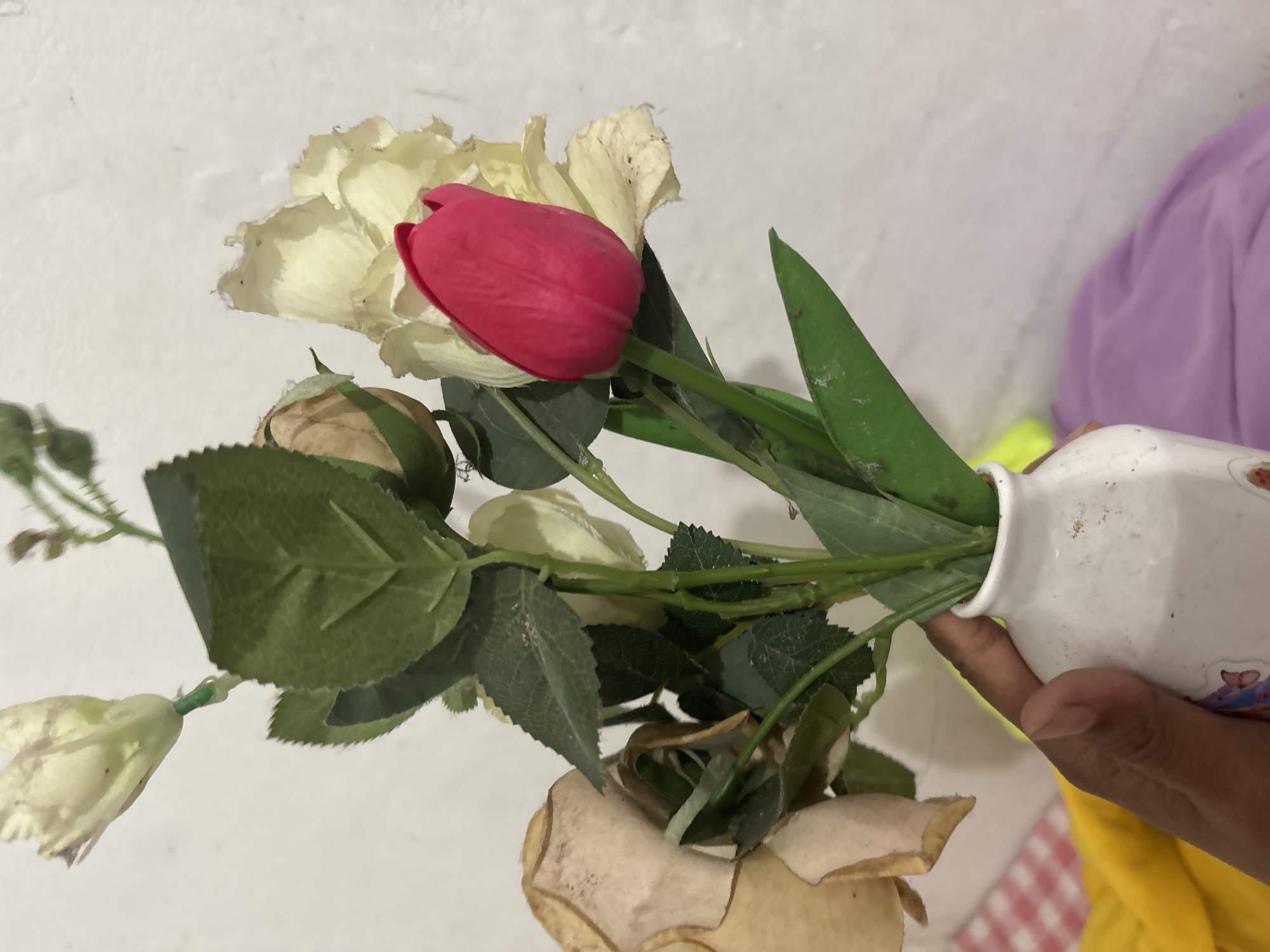 A hand is holding a white vase containing a bouquet of artificial flowers. The bouquet includes several white roses, one of which is wilted and brown, and a single red tulip bud. The background is a plain, textured white wall. The lower portion of the image shows a colorful arrangement of fabrics, including a red and white checkered pattern, a bright yellow fabric, and a purple fabric. The scene appears to be indoors, likely in a residential setting in North Jakarta, Indonesia, as indicated by the context.