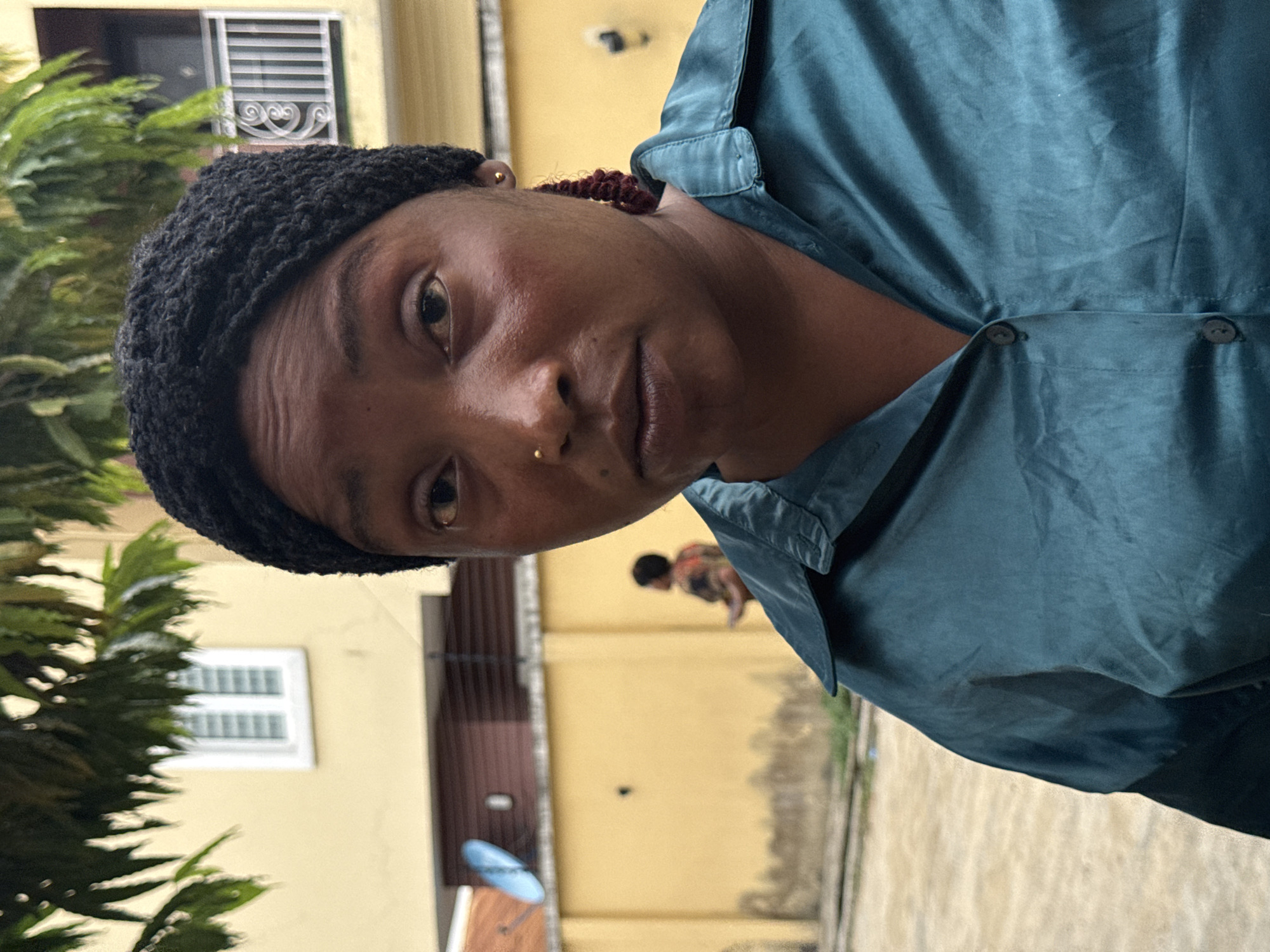 A medium shot features a dark-skinned individual wearing a black crocheted head covering and a shiny teal collared shirt. The person has a nose piercing. In the background, a yellow building with a white window is visible to the left, and a person in dark clothing walks in the distance to the right. The location appears to be outdoors in Ado, Nigeria, based on the style of the buildings and vegetation. A satellite dish is partially visible in the background.
