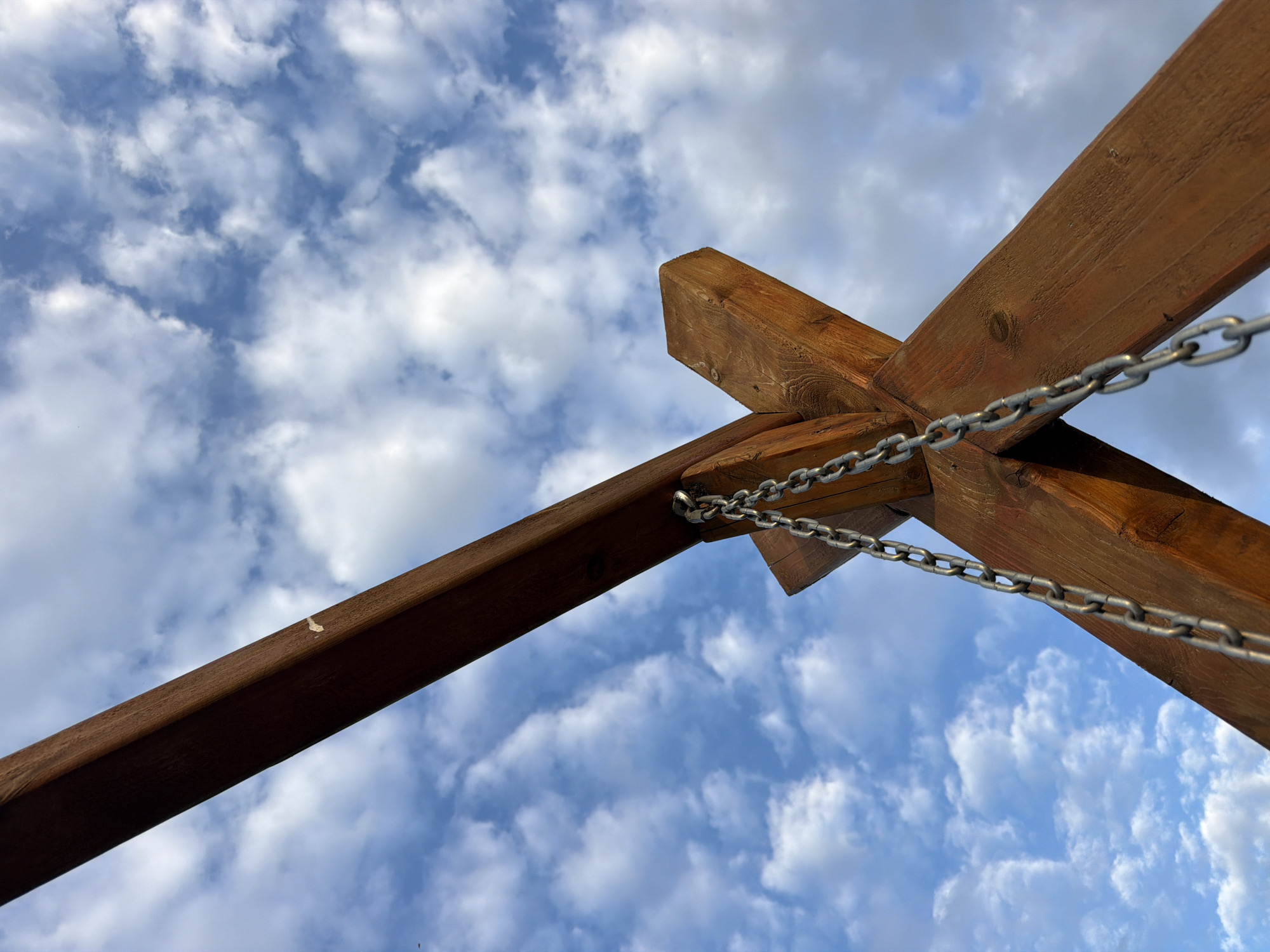 The image shows a close-up, low-angle view of wooden beams forming a cross shape, against a backdrop of a partly cloudy sky. Metal chains are attached to the beams, suggesting they are part of a structure, possibly a swing set or some kind of outdoor play equipment. The wood appears stained or treated, with visible grain and some knotting. The sky is a vibrant blue, dotted with fluffy white clouds, indicating fair weather. The lighting suggests it could be daytime, possibly late afternoon given the slightly diffused light and the angle of the sun not being directly overhead. There are no people visible in the frame, and the focus is on the construction and the sky. The overall impression is one of outdoor leisure or a partially constructed outdoor feature. The location context is Bol'shaya Ven'ya, Russia. There is no visible text in the image.