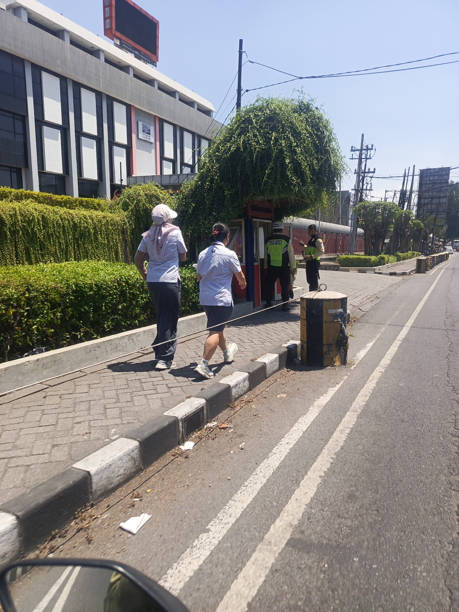 The image shows a street scene in Bungurasih, Indonesia. Two individuals are walking on a paved sidewalk beside a street. The person in the foreground on the left is wearing a light-colored shirt, dark pants, and a headscarf. The second person, walking slightly ahead and to the right, is wearing a light-colored shirt and dark shorts. Behind them, near a small kiosk or booth, are two individuals in what appear to be security uniforms, one wearing a bright green vest with "SATPAM" written on it, and the other a yellow vest. The environment includes lush green hedges bordering the sidewalk, a building with a modern facade and large windows in the background, and utility poles with electrical wires overhead. The street itself has clear white lane markings and a yellow and black striped bollard on the side. The sky is clear and blue, indicating a sunny day.