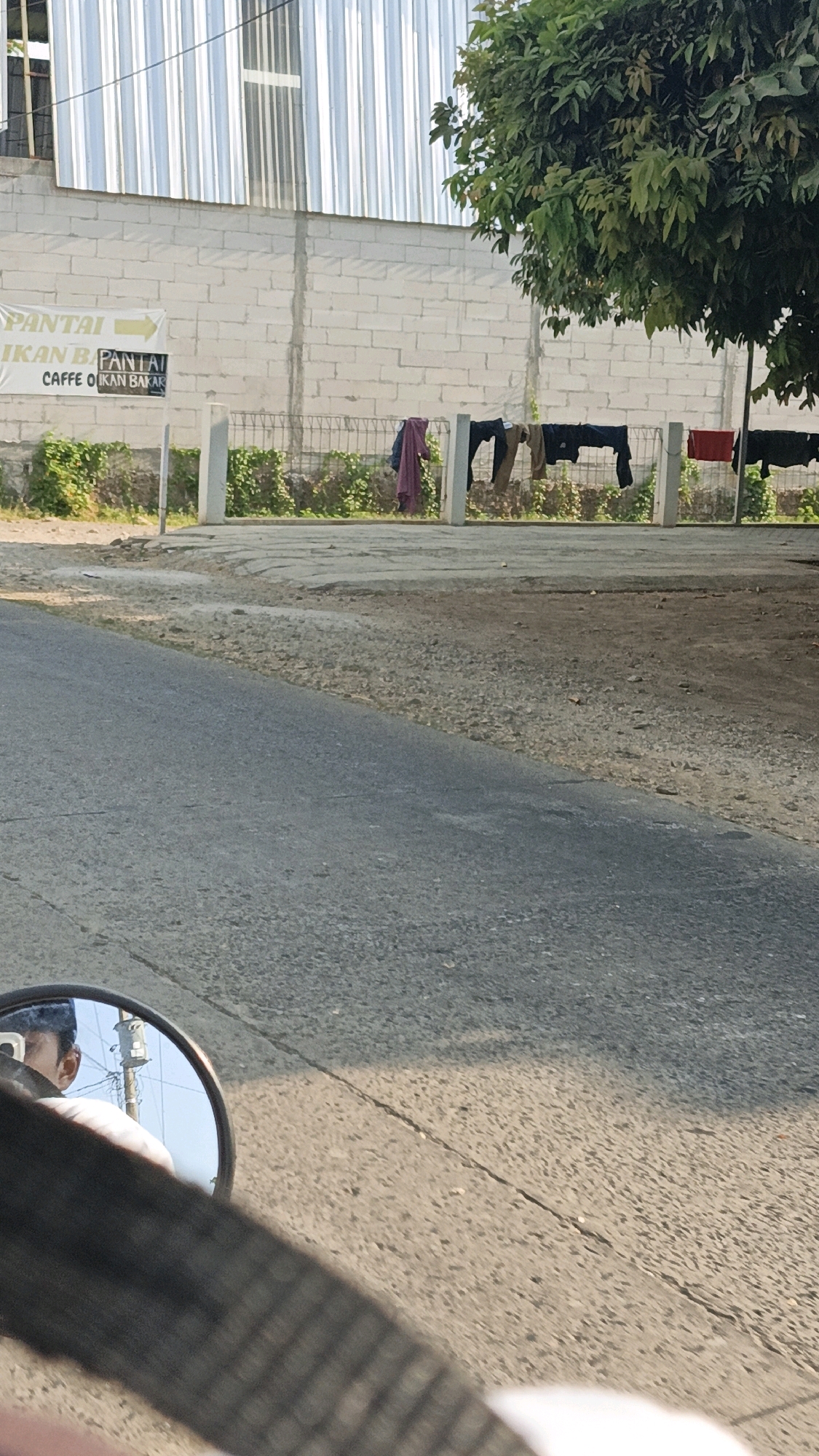 In Jepara, Indonesia, a low-angle shot captures a roadside scene. In the foreground, a motorcycle's rearview mirror reflects a person's head and shoulders, suggesting the viewer is riding a motorcycle. The road surface is asphalt, with patches of gravel and sand on the shoulder.
Behind the road, a white brick wall runs horizontally, topped by a metal-clad building with vertical corrugated panels. A fence with metal bars and white posts separates the road area from the wall. Clothes, including a red garment and dark-colored items, are hung on a clothesline stretched between the fence posts. Green foliage is visible growing along the fence and behind it. A sign board with Indonesian text, including "PANTAI" and "IKAN BAKAR" (grilled fish), is posted near the wall. A large tree with dense green leaves partially obscures the top right portion of the scene. The lighting suggests it is daytime, with sunlight casting shadows.