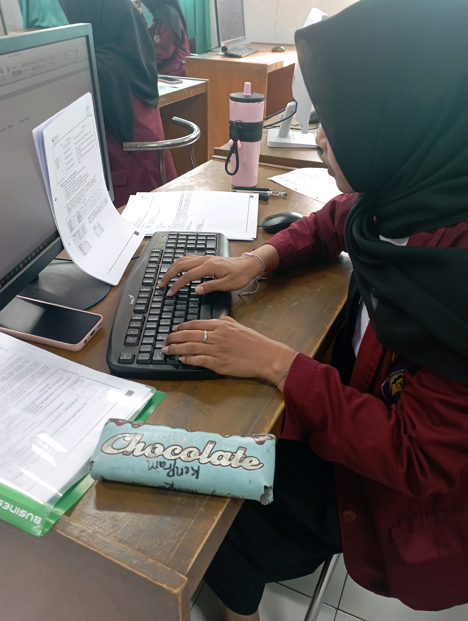 The image depicts a young woman wearing a black hijab and a maroon jacket, seated at a wooden desk in what appears to be a classroom or office setting. She is actively typing on a black keyboard. To her left, a computer monitor is visible, displaying a spreadsheet. A pink smartphone lies on the desk in front of the monitor. Several documents are spread across the desk, including a stapled packet on the left and loose papers to the right. A light blue object with "Chocolate" and "Kenji" written on it is positioned in the foreground. A pink water bottle with a black strap is also on the desk. In the background, another person wearing a similar maroon outfit and hijab is partially visible. The environment suggests an educational or administrative facility, likely located in Kawali, Indonesia, based on the prompt.