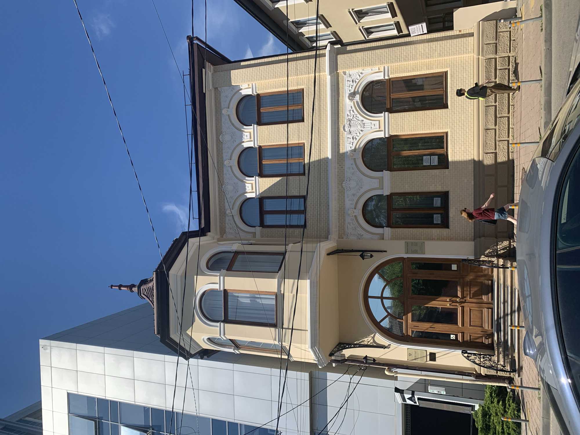 The image displays an urban street scene on a bright, sunny day, featuring two contrasting buildings, people, and a parked car. The dominant structure is an ornate, light yellow or cream-colored building with decorative white stucco elements. It features multiple arched windows with dark frames, some with drawn curtains, and a large, arched wooden main entrance door with glass panels. A decorative finial adorns the building's roofline.
To the left of this historical building, a modern structure with white/light gray square paneling and large glass windows is partially visible, creating a stark architectural contrast. Overhead, numerous black power lines crisscross the clear blue sky, which has a few wispy clouds.
On the sidewalk in front of the ornate building, strong shadows are cast by poles and the building itself, indicating direct sunlight, likely mid-morning or afternoon. Two people are present: one person, wearing a dark vest over a light shirt and dark pants, stands near a pole on the right side of the sidewalk. Another person, wearing a dark top and shorts, is partially visible walking on the far right, obscured by a silver car parked along the curb. Several light-colored bollards with yellow tops line the edge of the sidewalk. Green bushes are visible near the base of the modern building and reflected in some windows of the ornate building.
Visible text on a plaque near the main entrance of the ornate building is difficult to read clearly, but one plaque appears to have a large word starting with "АРХИВЫ" (Archives) and another below it with "МВК".