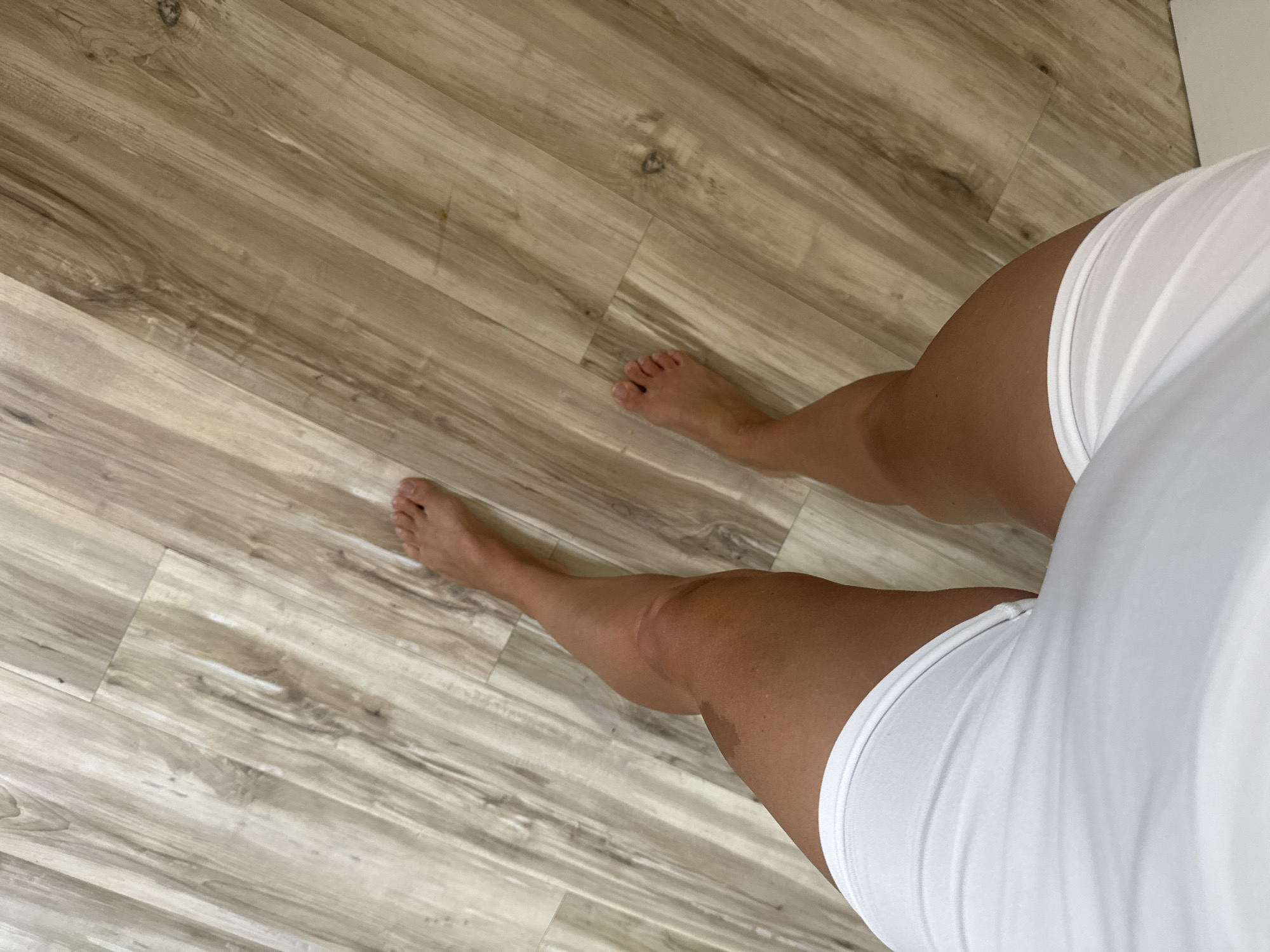 This is a high-angle, close-up shot of a person's legs and feet standing on a wooden floor. The floor has a light brown, wood-grain pattern with visible lines and textures. The person is wearing white underwear or shorts. The legs are tanned, and there is a darker mark on the left thigh. The scene is indoors, on a flat surface. There is no indication of the specific location within Baan Chaweng, Thailand.