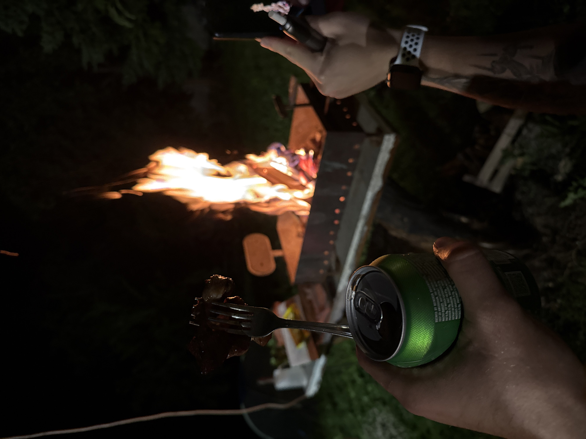 The image depicts a nighttime outdoor scene in Pet Kladentsi, Bulgaria, centered around a lit campfire contained within a rectangular metal brazier. In the foreground, a left hand holds a green aluminum beverage can, an open-top Heineken-branded container with visible condensation, and a silver metal fork piercing multiple pieces of dark, cooked meat. To the right, a second hand holds a dark cylindrical vape pen or e-cigarette. This right hand wears an Apple Watch with a white band, and a tattoo is visible on the forearm. The fire is brightly burning within the brazier, which rests on an elevated light-colored platform. The background is dark and out of focus, showing indistinct trees and shadowy ground cover, with some blurry white objects, possibly refuse or serving items, visible near the fire pit.