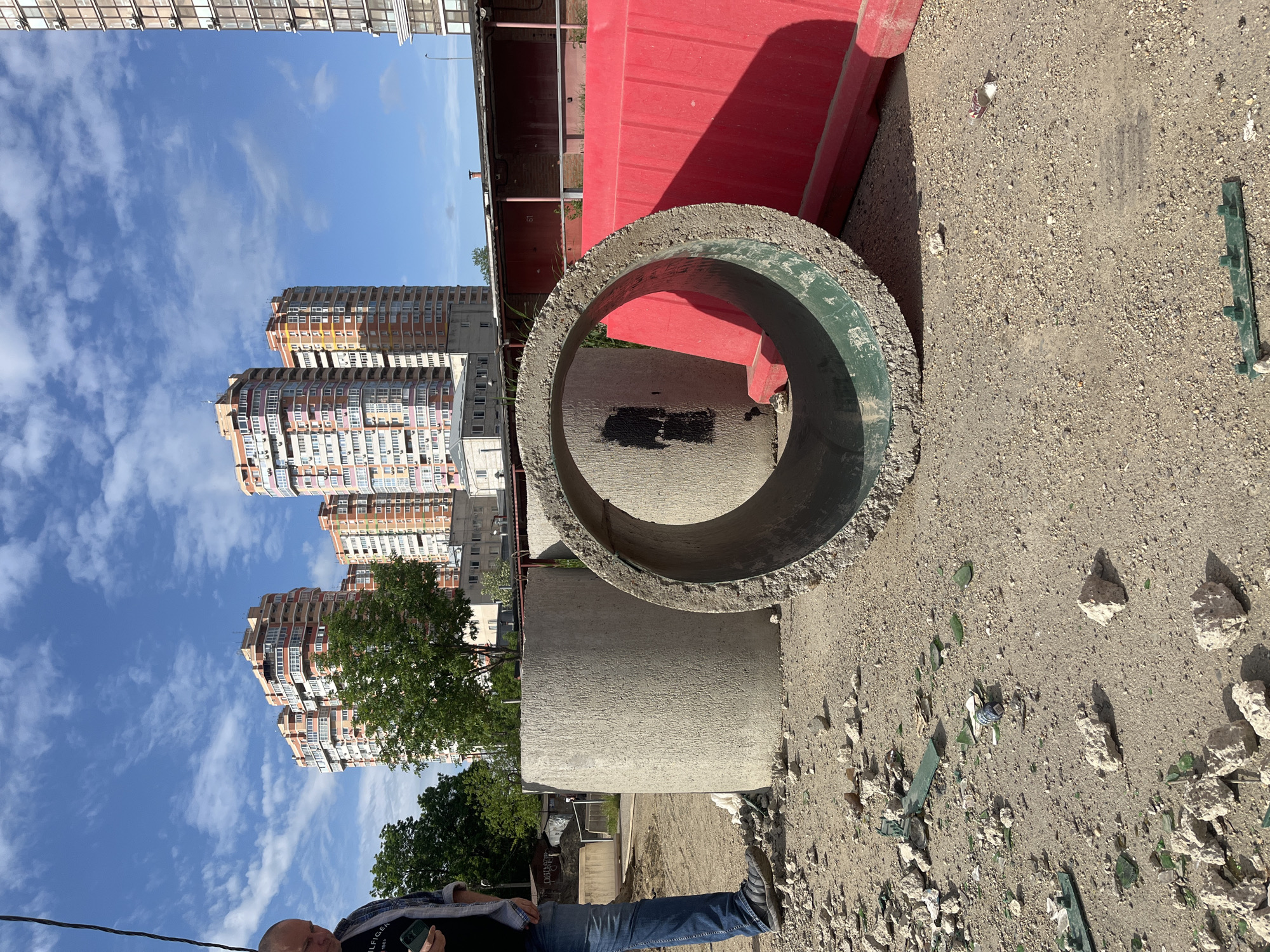 The image presents an outdoor scene in Kozet, Russia. The sky is blue with scattered clouds, and buildings are visible in the background.
In the foreground, a large concrete ring lies on a dirt surface strewn with rocks and shards of glass. A red barrier is situated next to the concrete ring. A man wearing a plaid shirt and jeans is partially visible in the bottom left corner of the image. He is holding a phone.
The scene suggests a construction or demolition site, evidenced by the concrete ring, barrier, and debris. The buildings in the background indicate an urban environment.
