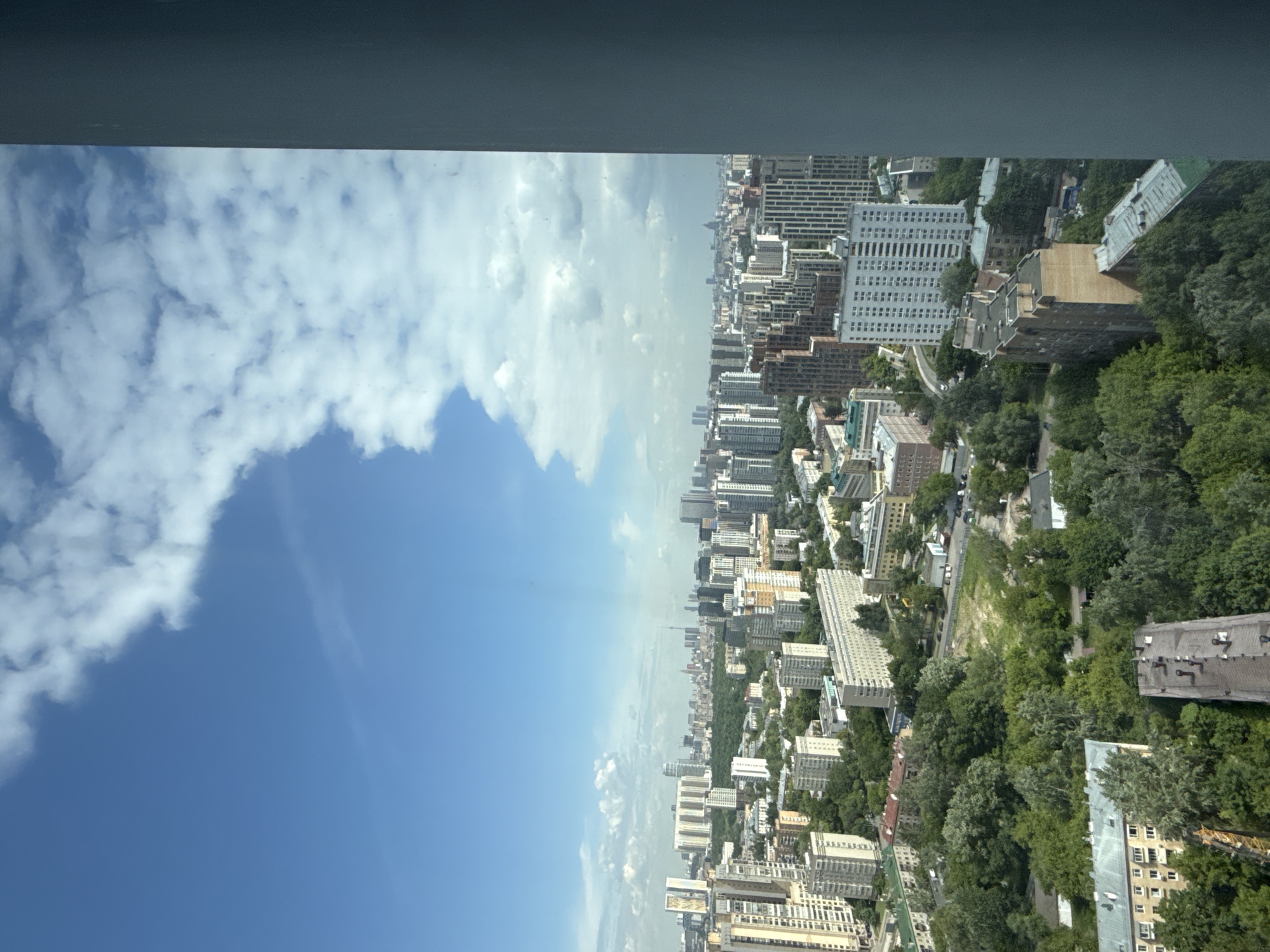 The image presents a high-angle, panoramic view of a sprawling urban landscape under a bright sky. The perspective is from an elevated position, looking out over a city. A dark, featureless band at the very top of the frame suggests an interior architectural element, likely part of a window frame or ceiling from which the photograph was taken.
The upper portion of the scene is dominated by a clear blue sky, interspersed with fluffy white cumulus clouds. Below the horizon line, the city extends far into the distance. It is densely populated with numerous multi-story buildings, appearing to be a mix of residential and commercial structures. Many of these buildings are rectangular and light-colored, typical of urban development.
Significant green spaces, including parks and tree-lined areas, are visible throughout the city, providing a contrast to the concrete structures. Roads and pathways can be seen winding through the urban fabric. The illumination suggests it is daytime, with clear weather conditions. While no specific landmarks are clearly identifiable, the overall appearance is consistent with a modern European or Russian city. Given the provided context, this is a view of Khimki, Russia.
There are no discernible activities involving people, and no visible text within the image. The scene conveys the vastness and density of an urban environment, balanced with natural elements.
