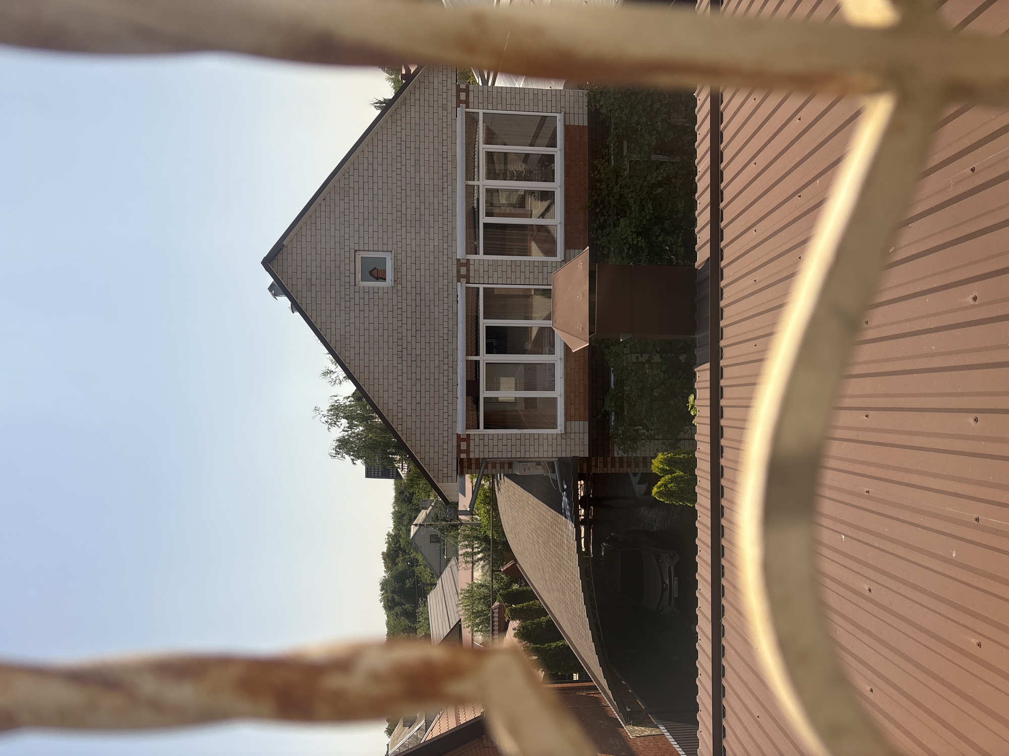The image shows a view from a window, looking out at a residential area under a clear blue sky. In the foreground, the blurry outlines of metal bars or a fence are visible, suggesting the vantage point is from behind a barrier.
Dominating the mid-ground is a two-story house constructed with light-colored brick. The roof is gabled and dark, with a prominent chimney. There is a smaller, square window on the upper gable where a person's face is visible looking outwards. Below this, on the first floor, are multiple large windows with white frames, reflecting the surrounding environment. A dark brown, possibly metal, structure is situated in front of the first-floor windows, resembling a small shed or air conditioning unit. Greenery, including ivy and some bushes, climbs the lower part of the house.
To the left of the house, a covered carport with a brown, textured roof shelters a car. The car appears to be a silver SUV with a license plate that is partially visible. Further left, more houses and rooftops are visible, along with numerous trees and dense foliage, indicating a suburban or semi-rural setting. The overall impression is of a sunny day in a quiet neighborhood.
The specific location is identified as the city of Georgiyevsk, Russia.