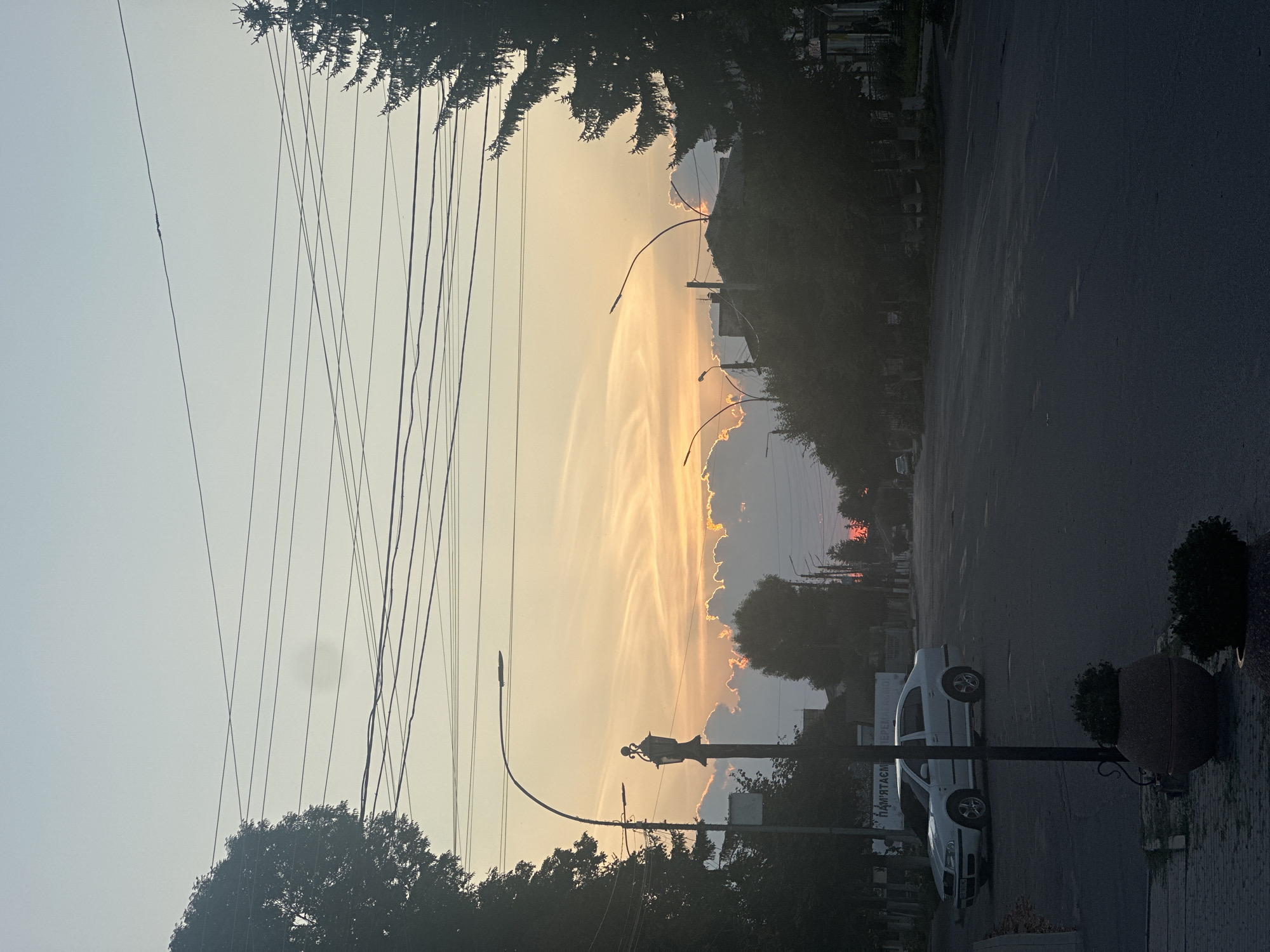 This is a street-level, full-shot photograph of a street in Khmilnyk, Ukraine, during what appears to be sunset. The sky is dominated by a soft gradient of pale blue at the top transitioning to warm oranges and yellows, with streaks of light clouds. Several power lines are visible crisscrossing the sky. On the left side of the frame, a silhouette of a large tree is prominent. In the foreground, a white sedan is parked on the right side of the road. A lamppost stands near the parked car, featuring an ornate, older-style lamp at its top, in addition to a modern street light on a curved arm. Two large planter pots are visible on the left sidewalk, one with a plant. The road surface is asphalt and appears to be in moderate condition, with some visible cracks and patches. The right side of the street shows more trees and the silhouettes of buildings. A smaller, red sun is visible on the horizon in the distance. The overall lighting suggests late evening, with a subdued and atmospheric quality.