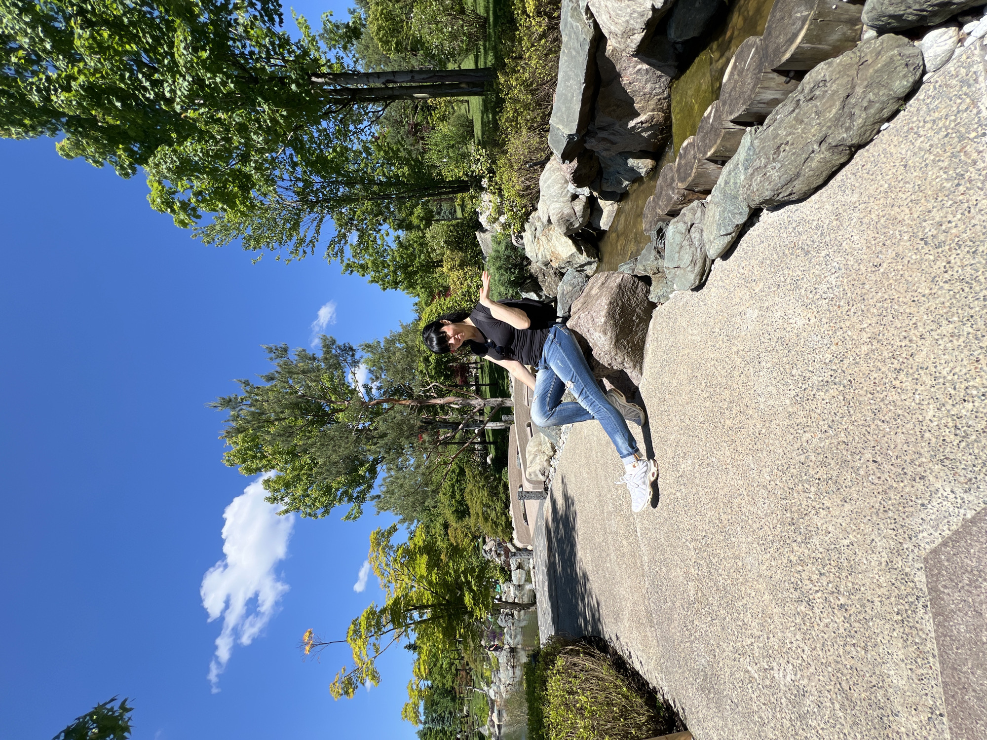 This media file captures a vibrant outdoor scene in what appears to be a landscaped park or garden. The image is taken on a sunny day with a clear blue sky dotted with a few white clouds.
In the mid-ground to the right, a woman with dark hair, wearing a black short-sleeved top, ripped blue jeans, and white sneakers, is posing. She is leaning casually against a formation of large rocks that line a narrow stream or water feature. Her right arm is raised in a gesture, and sunglasses rest on her head. The stream, visible to her right, flows gently between the rocks.
The foreground is dominated by a light-colored, pebbled concrete path where the woman stands. Further along the stream, wooden logs are incorporated into the rockwork, possibly forming a decorative element or part of a small bridge.
The background is rich with lush green foliage from numerous trees of varying heights and types, creating a dense canopy under the bright sunlight. Shadows are visible on the path and ground, indicating a strong light source, likely midday. The overall setting suggests a well-maintained public park or botanical garden, possibly designed with a Japanese garden aesthetic, located in Krasnodar, Russia. No visible text is present in the image.