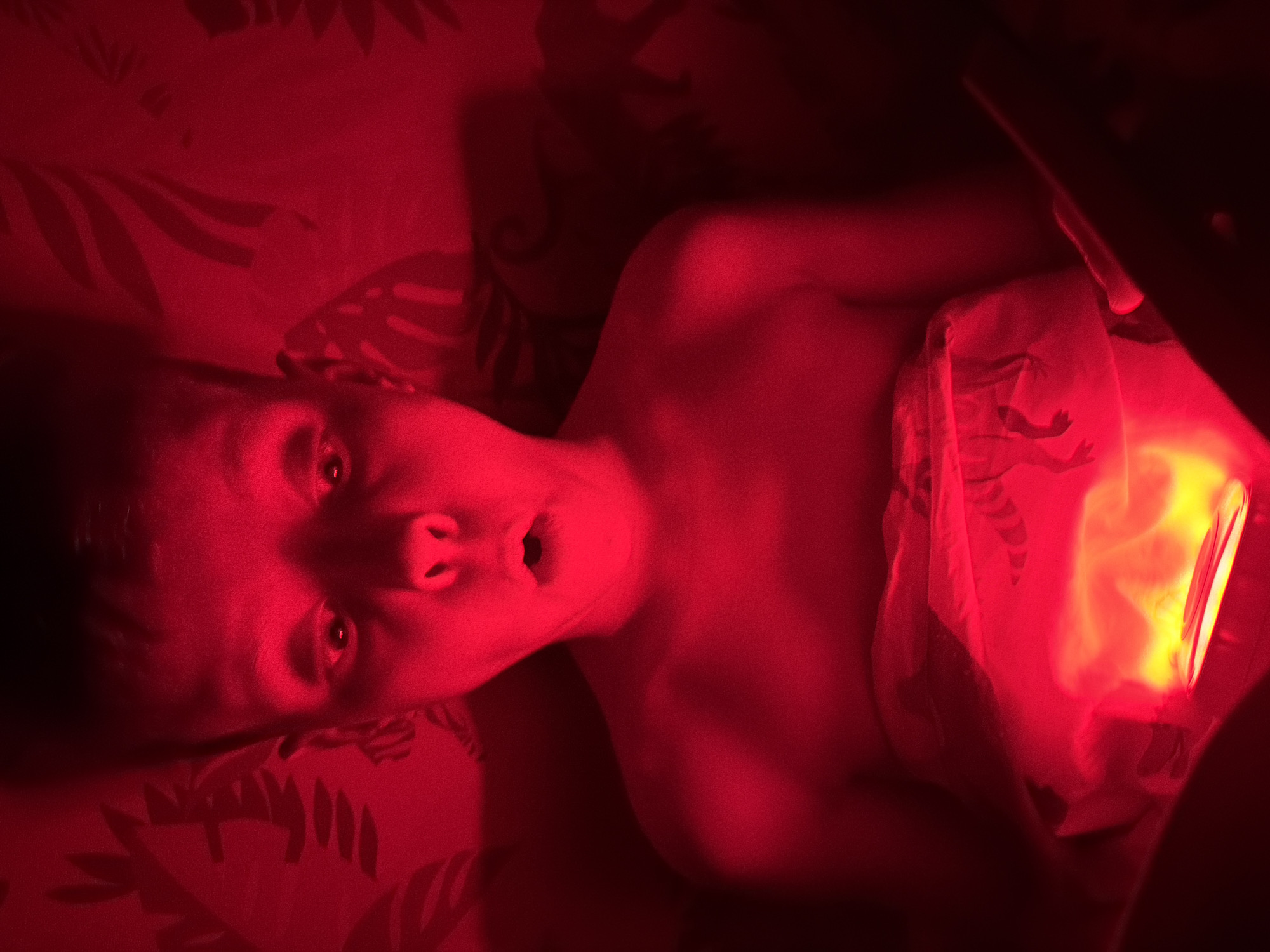A shirtless child lies on a patterned bed, illuminated by a red light. The child's expression is one of surprise or alarm, mouth open, eyes wide. The red light emanates from what appears to be a tablet or similar device, partially covered by a white blanket. The background suggests a bedroom environment. There is no verifiable information to suggest this photo was taken in Votkinsk, Russia.