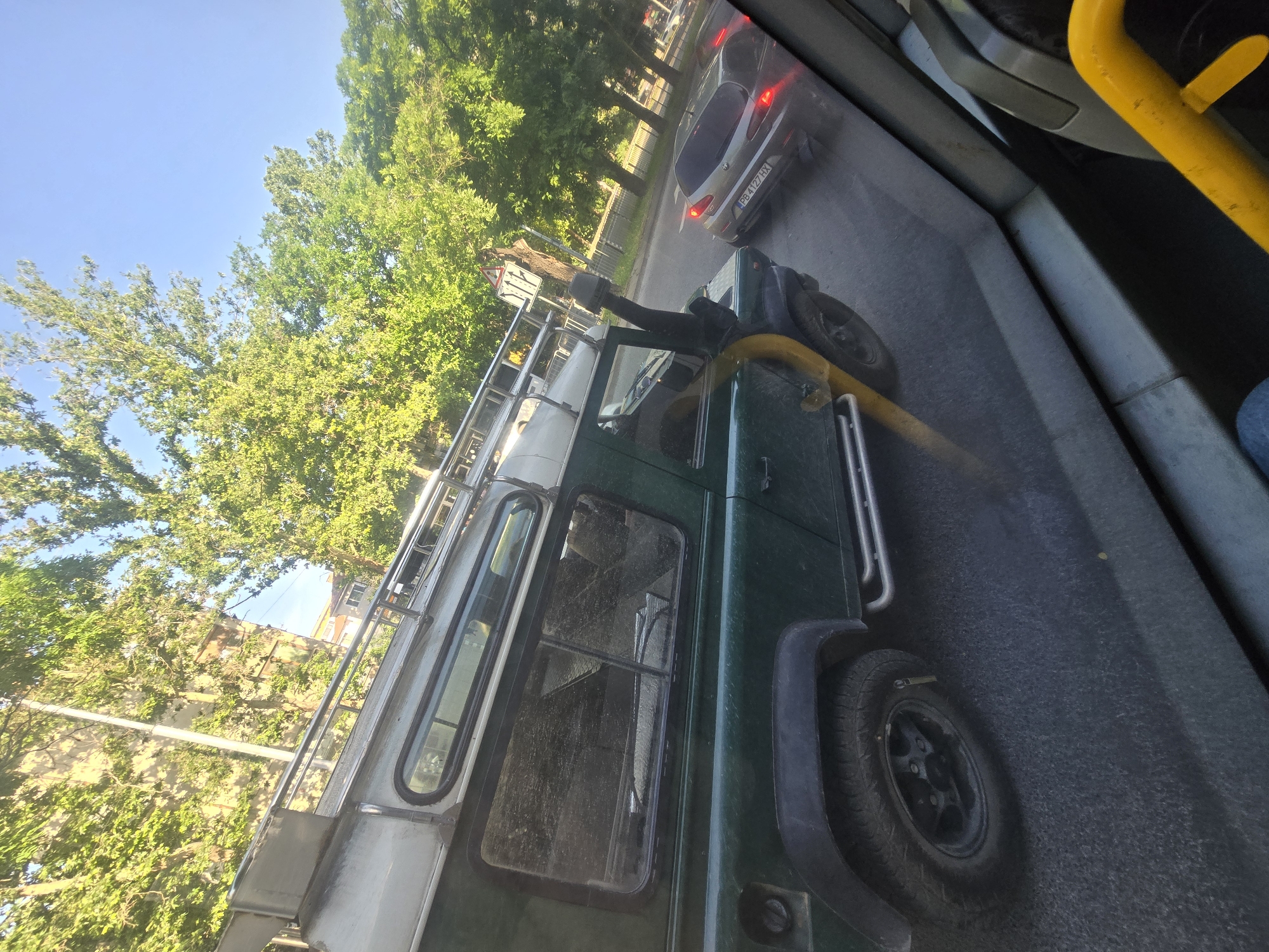 A medium shot depicts a green and silver Land Rover Defender with a snorkel and roof rack, viewed from the inside of a bus. The Defender is moving along a street, as suggested by the presence of a silver car with a license plate. The environment includes greenery and a building in the background, indicating an urban or suburban location. A yellow pole is visible inside the bus, and a person’s leg is visible on the right side of the image. The city and exact location remain unknown.