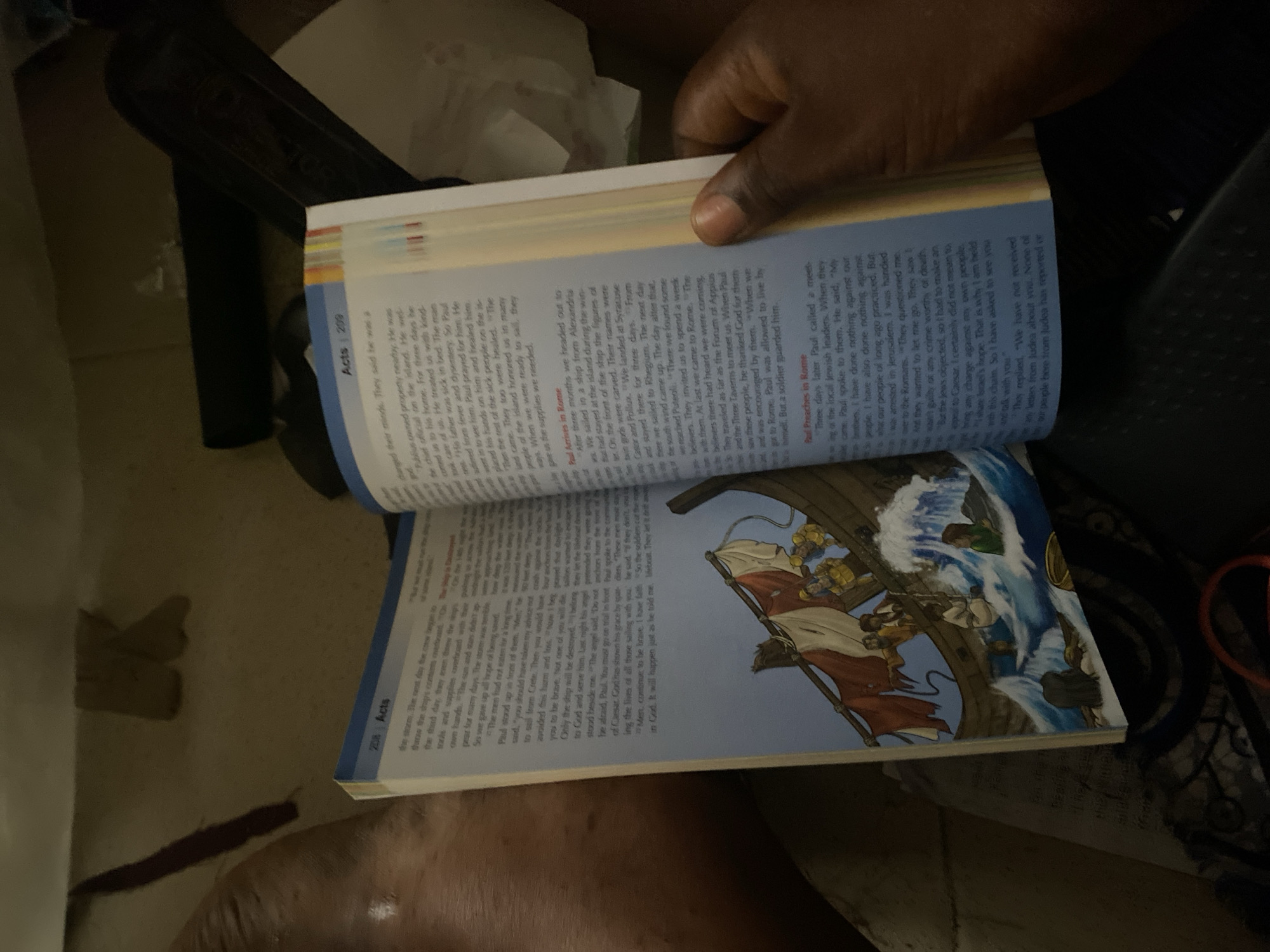 The image shows a person's hand holding an open book, with the left page displaying text and an illustration of a boat in choppy waters. The right page of the book continues with text. The book appears to be a religious text, likely the Bible, given the chapter title "Acts" visible on both pages. The illustration depicts a historical scene with people on a ship being tossed by waves. The background is dark and out of focus, with glimpses of what might be furniture or other objects. The lighting suggests an indoor setting. There is no discernible information in the image that would indicate a location in Jauro Yinu, Nigeria.