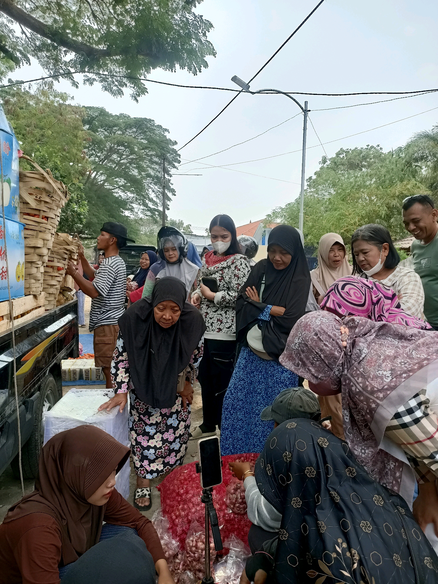 The image depicts an outdoor market scene in Madiun, Indonesia, where a group of people are gathered around a truck loaded with goods, possibly produce. A man in a striped shirt and shorts is seen handling baskets of items from the truck. Several women, some wearing hijabs, are looking at the goods or engaged in conversation. One woman wears a helmet and a face shield. Bags of onions are visible on the ground, suggesting a sale is in progress. The environment appears somewhat rural or suburban, with trees and power lines in the background. A cell phone on a tripod is positioned near the onions, potentially for recording or live streaming.