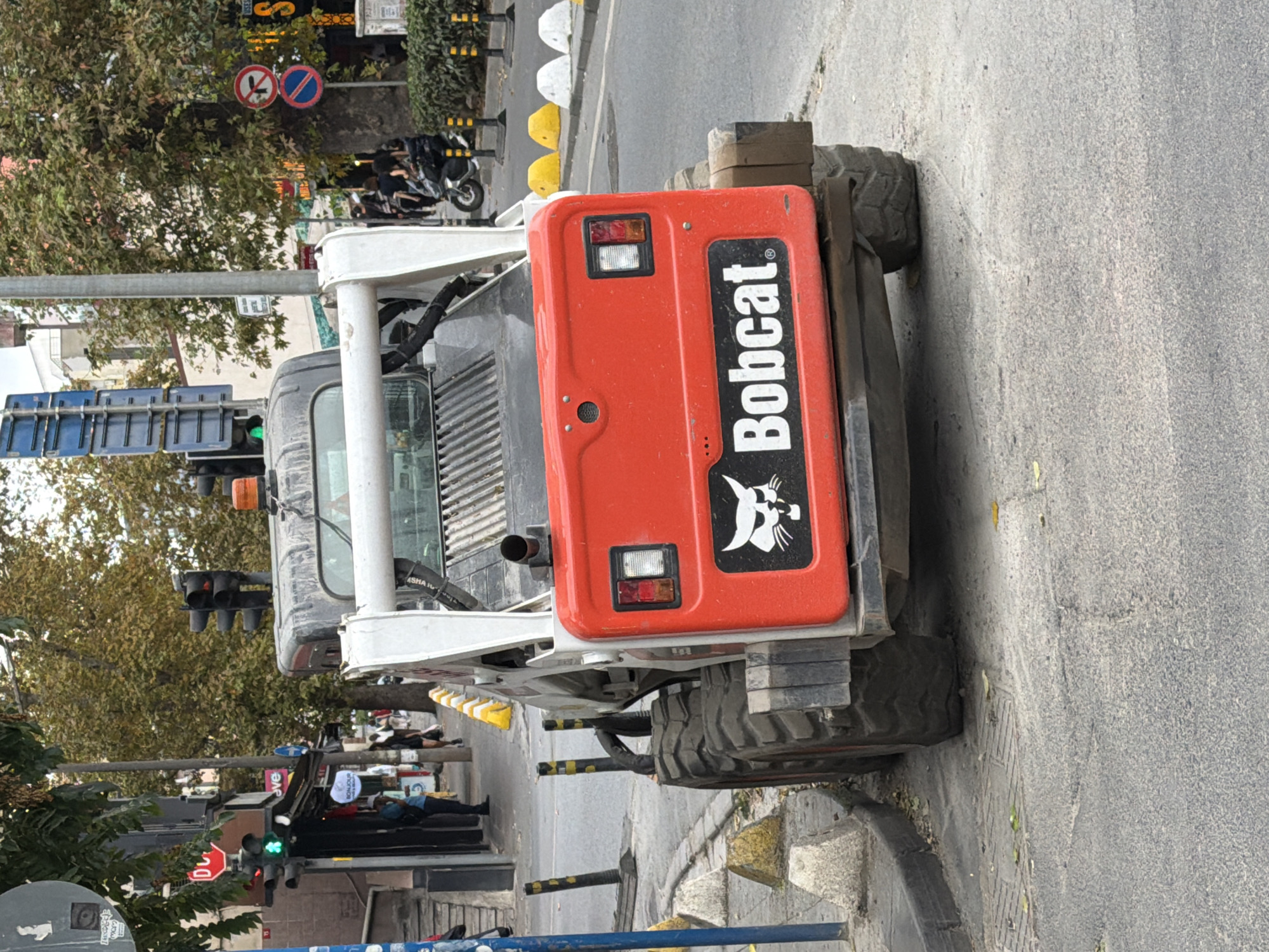 The image shows a Bobcat skid-steer loader parked on a street in Kadıköy, Turkey. The skid-steer loader is red and white with large, rugged tires. It is positioned at an angle on the asphalt road, close to a sidewalk. The background shows a street scene with trees, buildings, traffic lights, and several people walking or standing. A red stop sign and a traffic light showing green are visible on the left side of the image. Yellow and white bollards line the sidewalk. The overall impression is a normal urban street scene, possibly during daylight hours, with the skid-steer loader being an active piece of equipment in the area.
