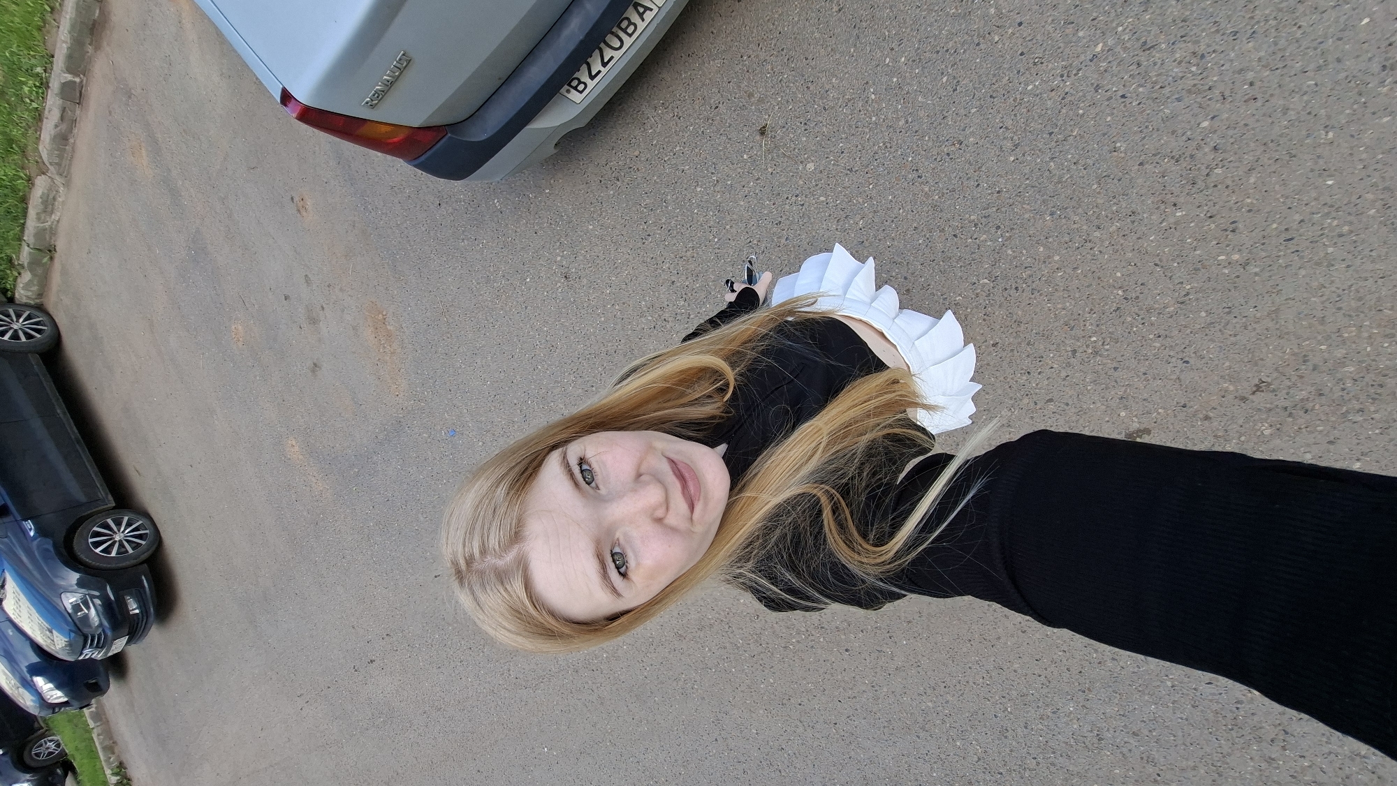 In the city of Chaikovsky, Russia, a young Caucasian woman with long blonde hair and green eyes is captured from a high-angle perspective, holding a smartphone to take a selfie. She is wearing a black long-sleeved top and a white pleated skirt. The background shows an asphalt-paved area, likely a parking lot, with several cars visible. To the right, a silver Renault car is parked, and its license plate displays the characters "BZZOBA". To the left, two blue cars are parked, one of which appears to be a Volkswagen Polo. The ground is a mix of asphalt and some dirt patches. The lighting suggests it is daytime, and the scene is outdoors.