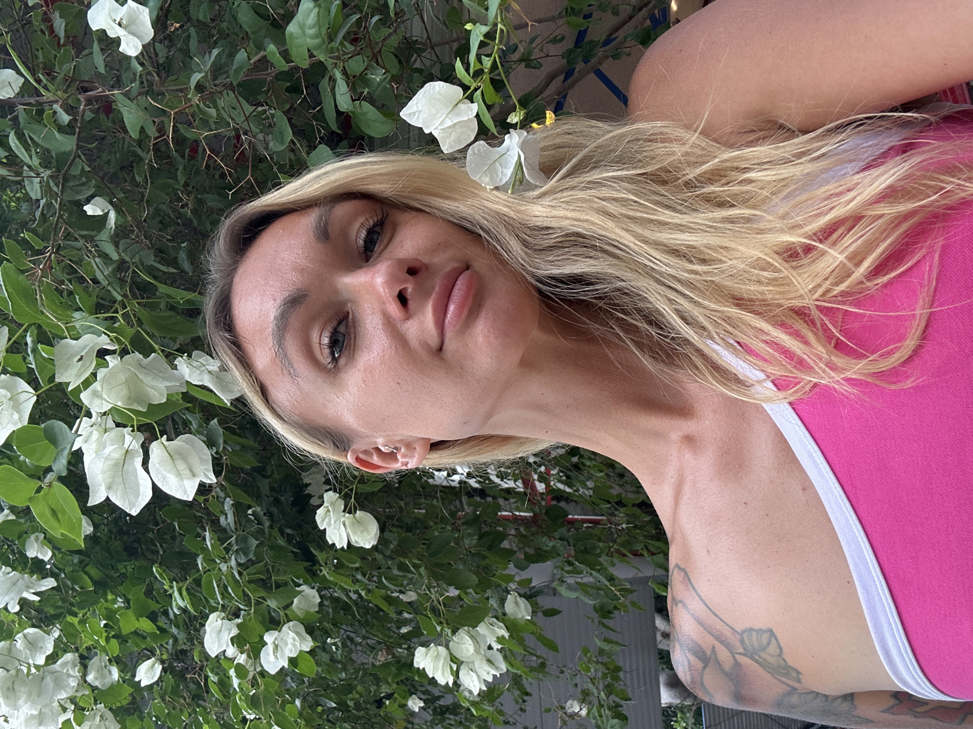 A woman with blonde hair and a pink top is in front of a wall of white flowers and green foliage. She is facing the camera and looking slightly to the left. She has a tattoo on her left shoulder. The background suggests an outdoor setting, possibly a garden or courtyard. The lighting is natural, indicating daytime. The image is a selfie. There is no indication of the location being in Phan Sơn, Vietnam.