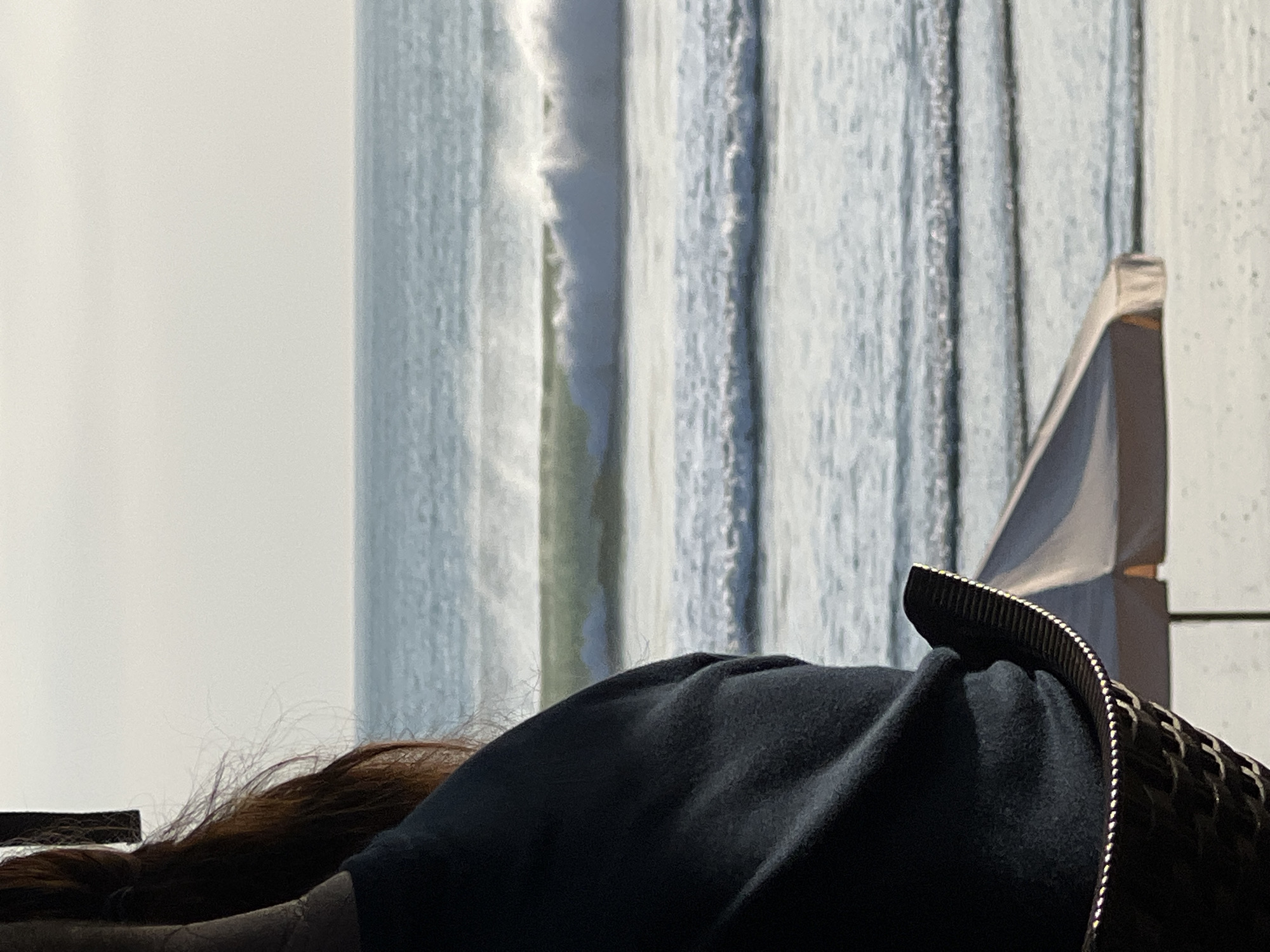 A person with auburn hair, wearing a dark blue shirt, is seated in a woven chair, looking out at the ocean. The person's back and side are visible in the foreground. The ocean features a series of waves, with a larger, breaking wave in the middle ground. The sky is overcast. In the lower right of the frame, the edge of a light-colored beach umbrella is visible. The scene is likely Kuta, Indonesia, a popular beach destination known for its surf.