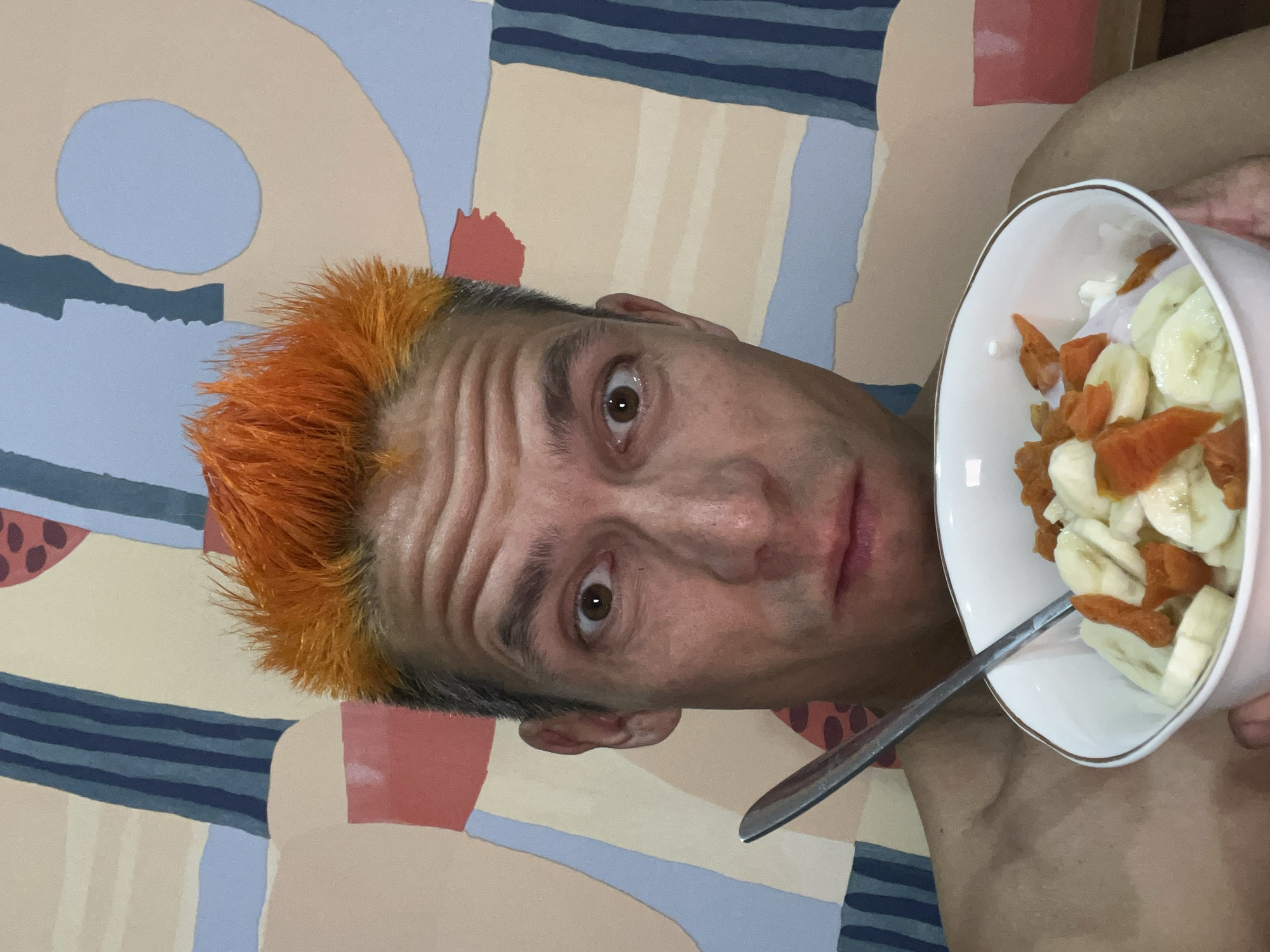 A close-up portrait shows a man with orange hair facing the camera and holding a bowl of sliced bananas and dried apricots. A metal spoon rests in the bowl. The background is a wall with a geometric, abstract painting. The image was taken in Chaikovsky, Russia.