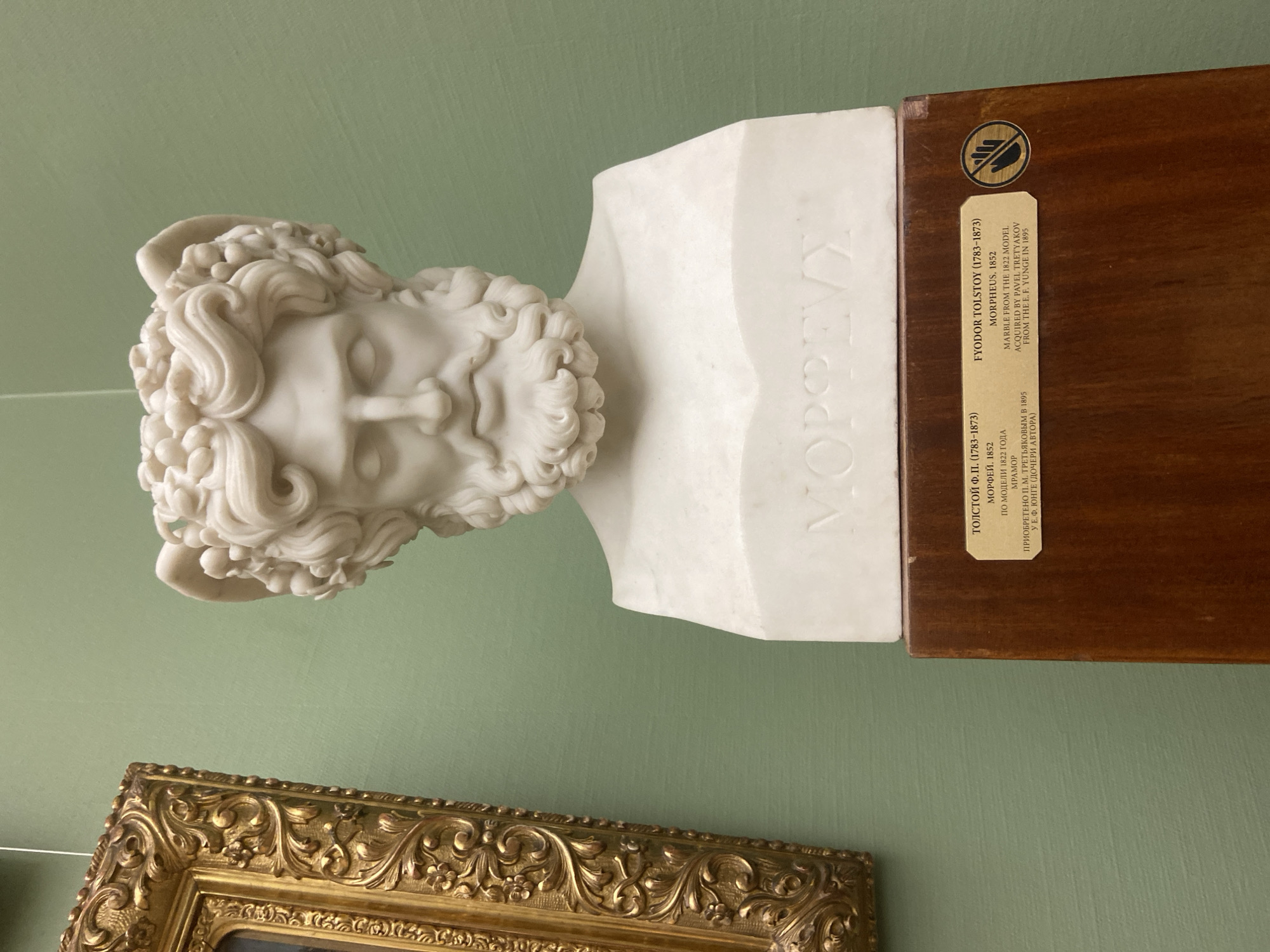 The image shows a marble bust of Morpheus by Fyodor Tolstoy, created in 1852. The bust is displayed on a wooden base with an informational label in both Russian and English. Below the bust, a decorative gold frame is partially visible. The bust is located in the city of Moscow, Russia.