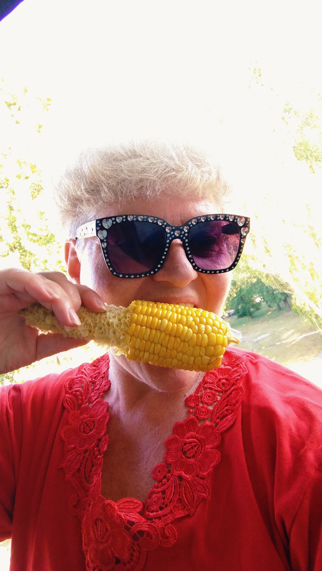In Horishni Plavni, Ukraine, a close-up shot captures a woman wearing decorative sunglasses and a red top with intricate lace trim. She is in the process of eating an ear of corn, holding it with her left hand and biting into it. The background is out of focus, showing greenery and sunlight, suggesting an outdoor setting.