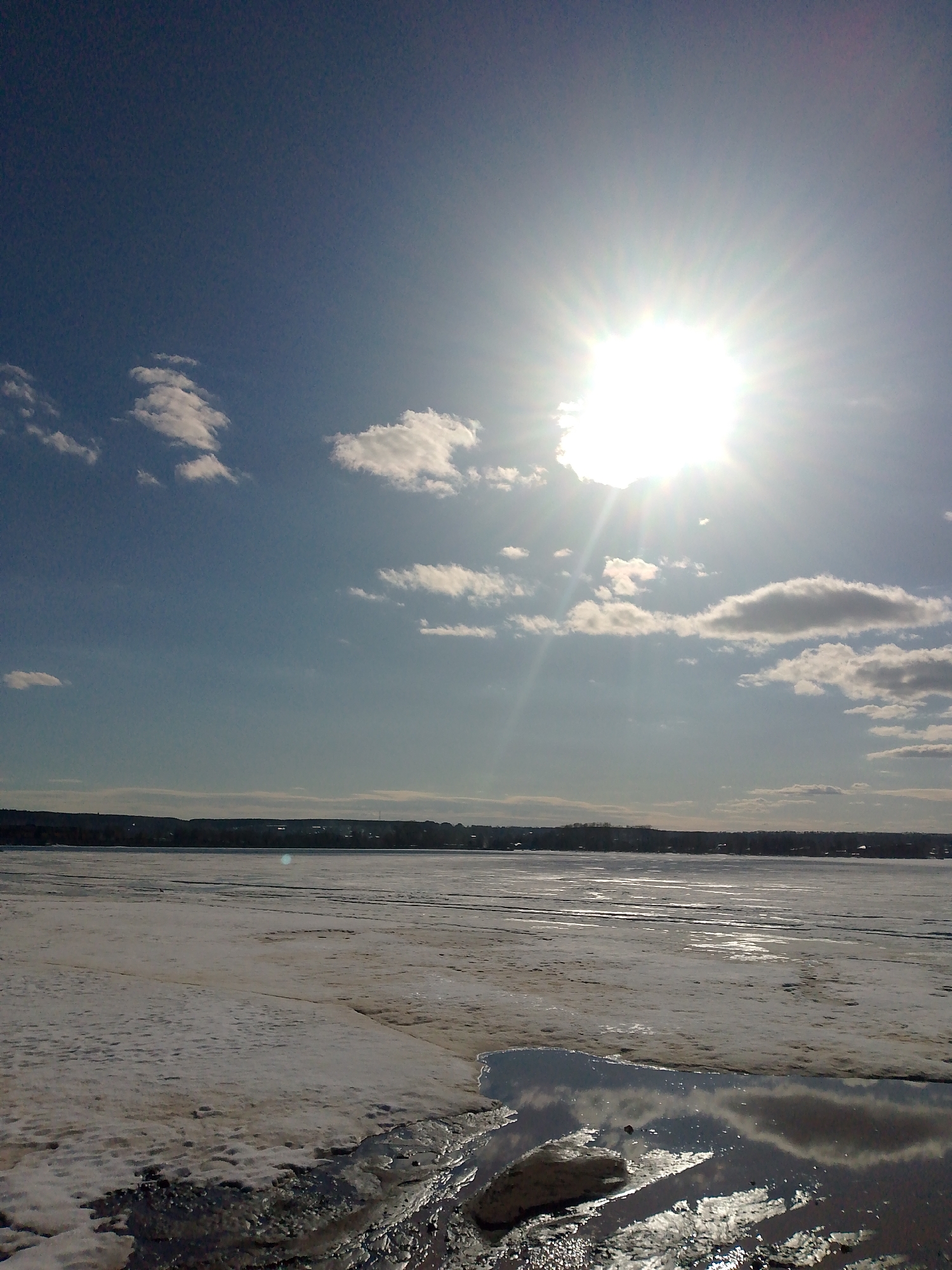 The image depicts a wintry scene in Votkinsk, Russia. A partially frozen body of water, likely the Votkinsk Reservoir or Kama River, dominates the foreground, with patches of snow and ice intermixed with open water reflecting the sky. In the background, a treeline and distant urban area suggest the outskirts of Votkinsk. Above, a bright sun shines through a partly cloudy sky, creating a high-contrast scene with the sunlight reflecting off the water and ice. There are no people visible in the image.