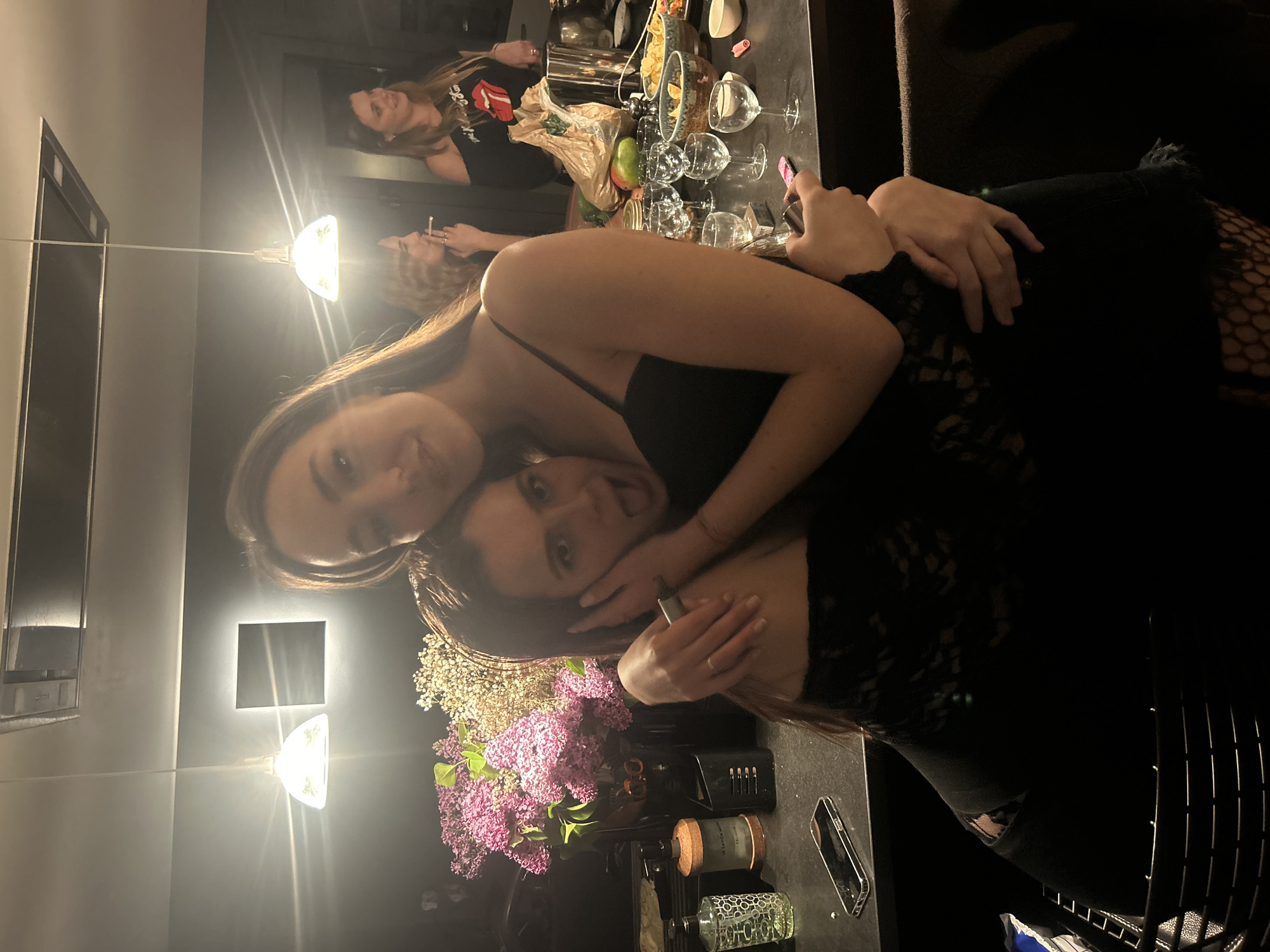 Two women are embracing indoors. The woman on top wears a black dress with spaghetti straps, while the woman underneath wears a black, lace-sleeved top and black jeans with fishnet stockings. They are positioned near a table holding various items, including flowers in a vase, candles, a phone, and personal grooming items. Other women are visible in the background. The location is in the city of Unknown, Unknown.