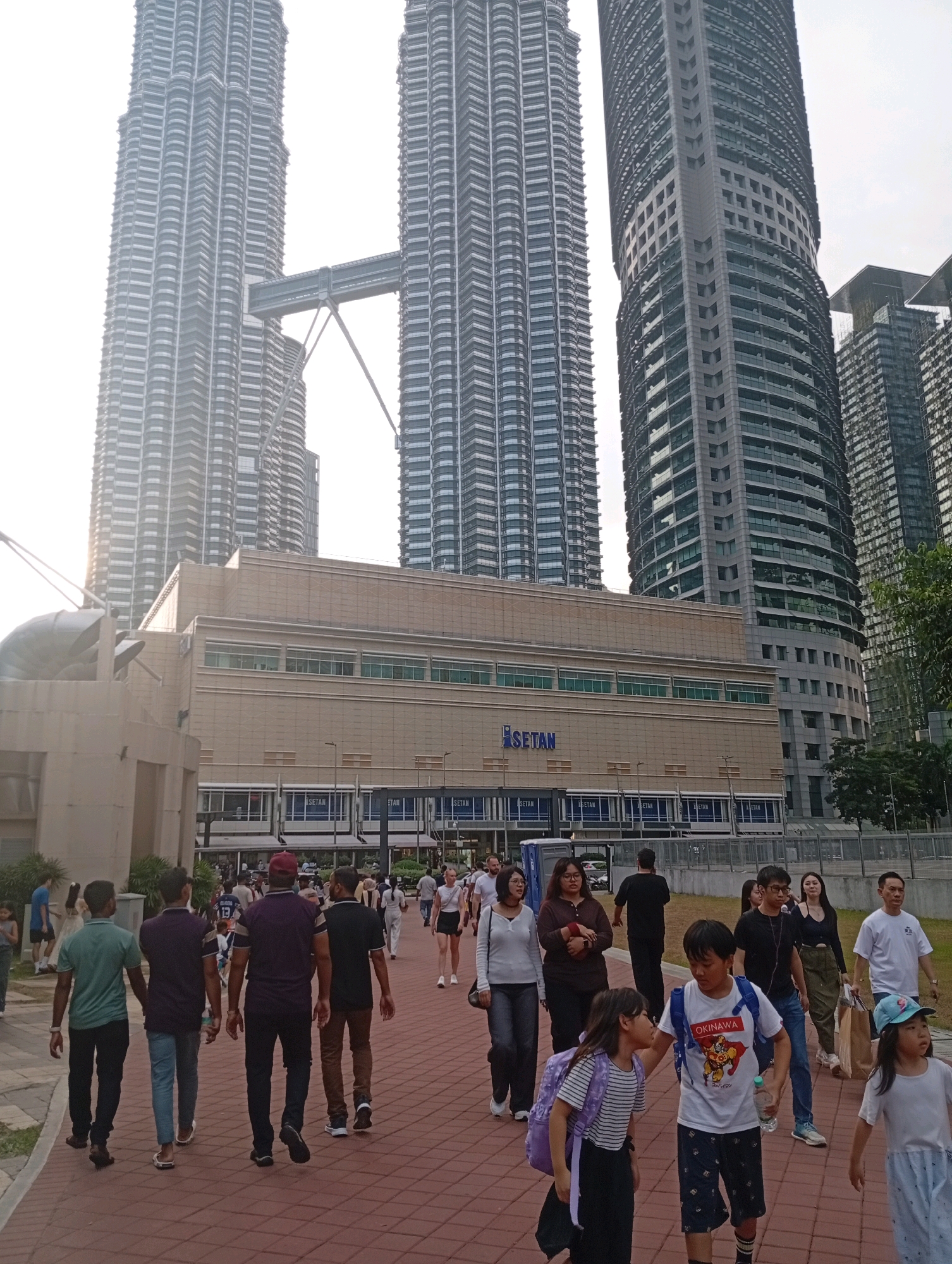 The image depicts a street scene in Kuala Lumpur, Malaysia, featuring the iconic Petronas Twin Towers in the background. The foreground is dominated by a paved walkway with numerous people walking in various directions.
In the middle ground, a modern building with the "SETAN" logo is visible, suggesting it is a commercial establishment or part of a larger complex. The architecture of the building is contemporary, with large glass windows.
The Petronas Twin Towers, a prominent landmark in Kuala Lumpur, rise majestically in the background. They are characterized by their distinctive twin spires and the skybridge connecting them. The sky is overcast, with a hazy quality to the light.
The people in the image are dressed in casual attire, suggesting a leisurely day. They are of diverse ages and ethnicities. Some individuals are walking alone, while others are in groups, indicating a public space with significant foot traffic. The overall atmosphere appears to be one of everyday urban activity.