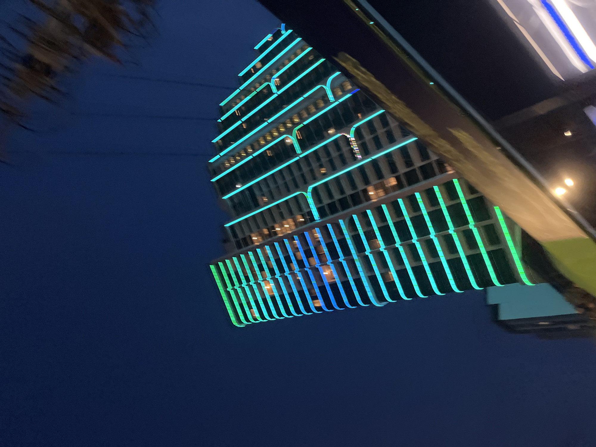 The image shows a modern, illuminated skyscraper at night in Nha Trang, Vietnam. The building's facade is outlined with vibrant green and blue lights that wrap around its curved edges and run vertically between floors. The lights create a striking visual contrast against the dark night sky. Individual windows on the building are lit from within, suggesting occupancy. In the foreground, a blurred, dark object, possibly a balcony railing or another structure, partially obstructs the view of the lower part of the building. The overall scene depicts an urban environment at night, characterized by architectural lighting and a sense of scale.
