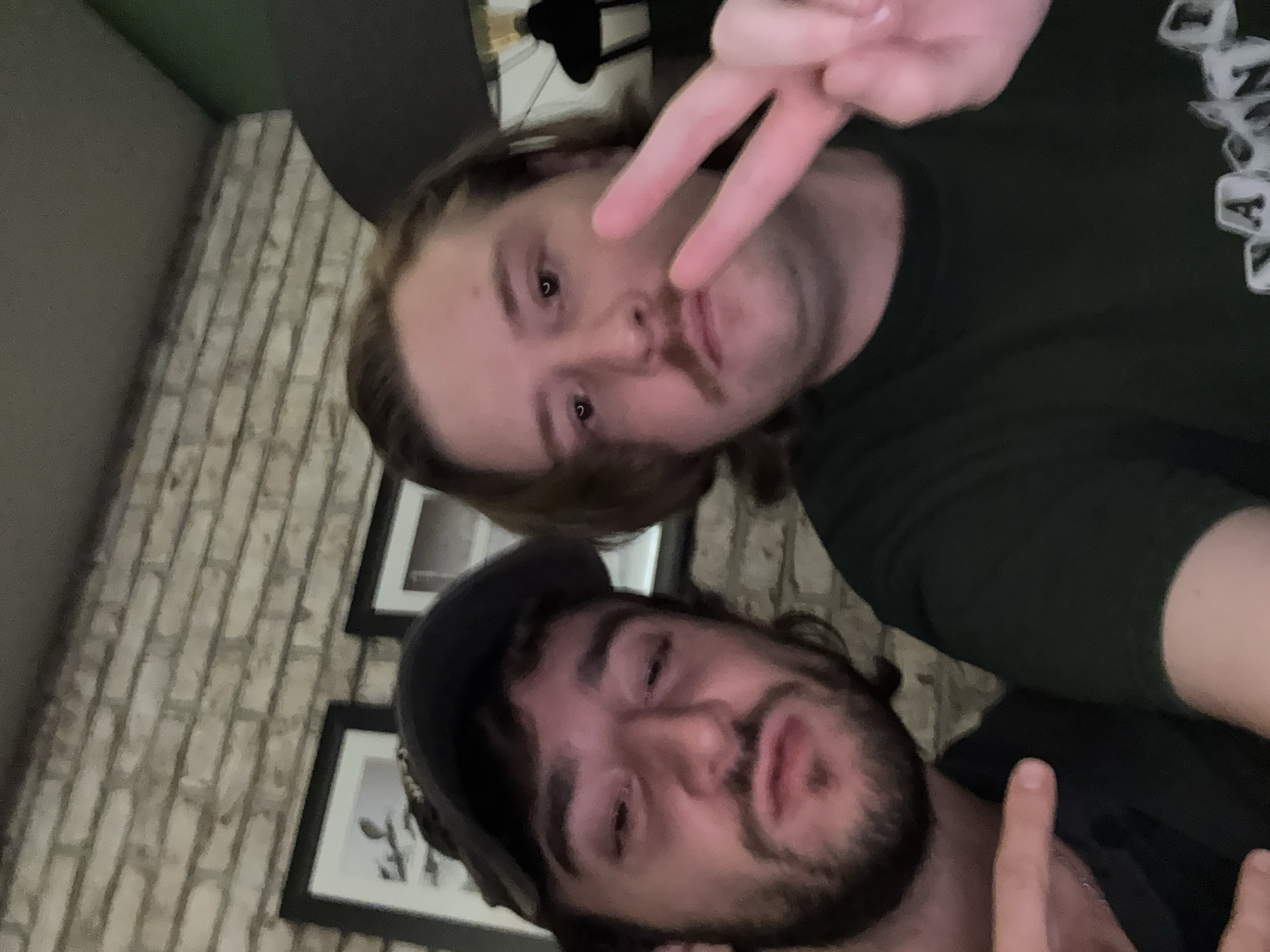 Two men are posing for a selfie. The man in the foreground has a beard and is wearing a baseball cap. The man in the background is making a peace sign. A brick wall and framed pictures are visible in the background. The location is possibly indoors, but the specific location in Huixquilucan de Degollado, Mexico, is unidentifiable from the image.