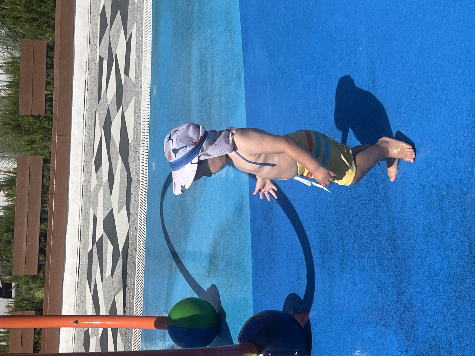 A young child wearing a sun hat and striped swim trunks stands on a blue, wet surface, likely a splash pad, in Sochi, Russia. The child is facing to the right, with their left arm slightly outstretched and their right hand raised. Two large, colorful spheres are partially visible to the left, attached to orange poles. The ground features a geometric pattern of grey, black, and white tiles leading to a white tiled edge. In the background, wooden benches and greenery are visible. The overall scene suggests an outdoor recreational area on a sunny day.