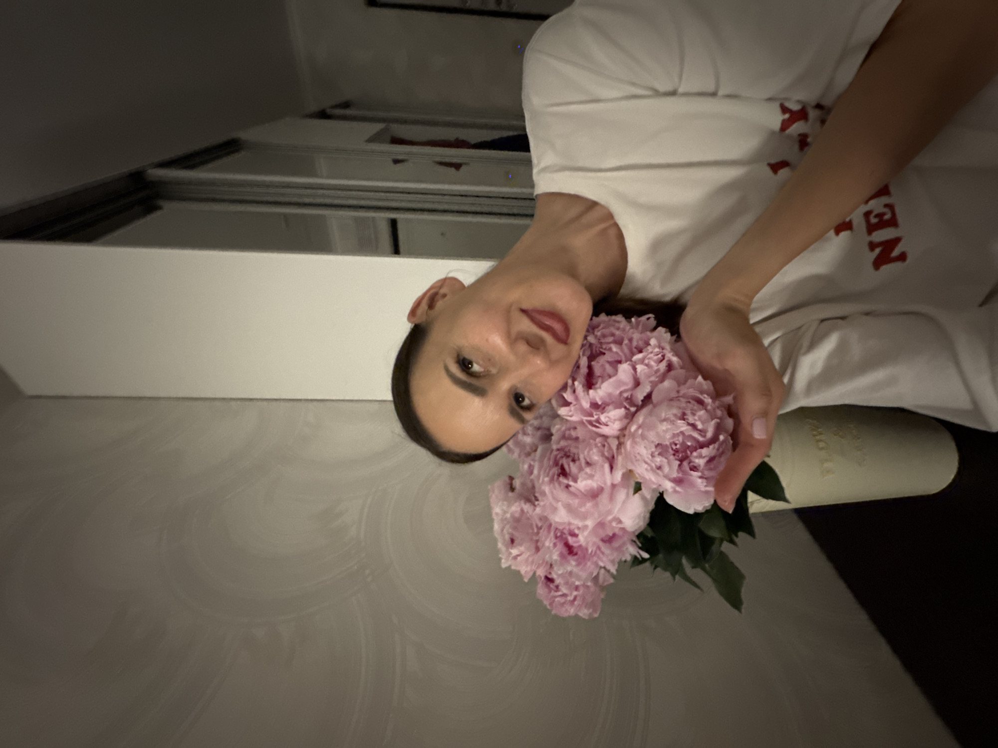 An adult woman, with brown hair pulled back and wearing a white t-shirt with red lettering that appears to say "NEW YORK", is captured in a medium shot, leaning her head against a bouquet of pink peonies. The woman is looking upwards and to her left with a slight smile, her right hand gently touching the flowers. The bouquet is in a light-colored vase placed on a dark surface. The background features a textured wall with a swirling pattern on the left and a closet with mirrored doors on the right. The lighting is dim, creating shadows. The image is devoid of any recognizable landmarks or indicators of the city of Sochi, Russia.