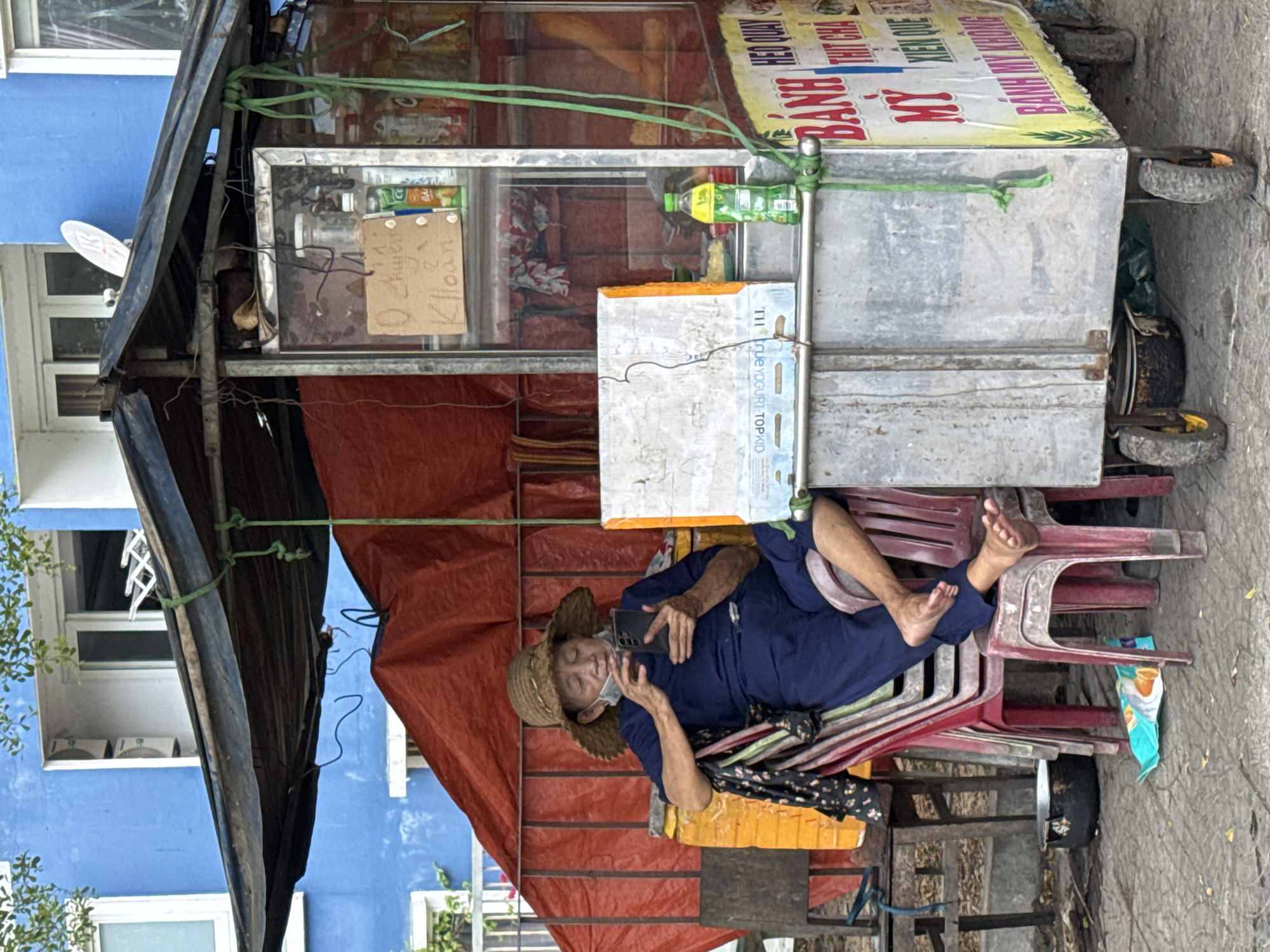 A rotated image shows an elderly Vietnamese woman resting in what appears to be a mobile street food vendor cart, likely in Đà Nẵng, Vietnam. The woman, wearing a straw hat, a surgical mask, and a blue outfit, is lying on a stack of red plastic chairs. She is holding a smartphone in her hands. The cart itself has a metal frame and a sign in Vietnamese. The surroundings include a blue building and a tarp.