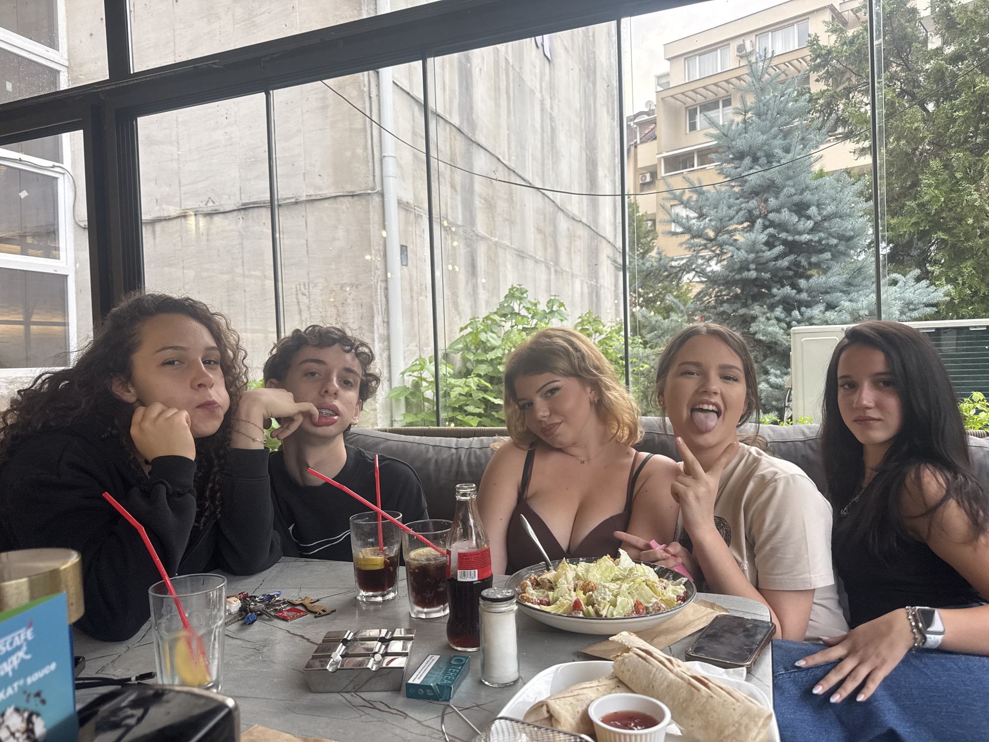 Five young people, three females and two males, are seated at a table on a patio. The table is covered with food and drinks, including a salad, wraps, soda, and a bottle of Coca-Cola. The individuals are interacting with each other, with some smiling and others looking at the camera. The background shows a cityscape with buildings and trees. The scene appears to be a casual gathering in a restaurant or cafe.