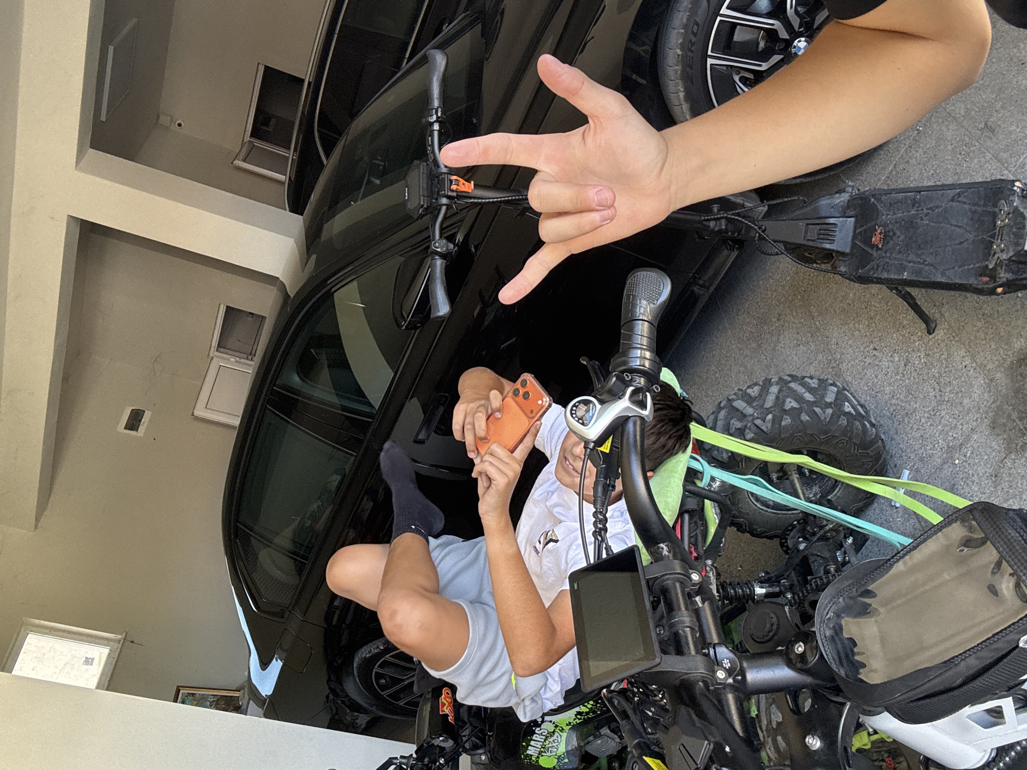 The image shows a young person seated on a black and green electric scooter within an enclosed garage. The person, wearing a white t-shirt, gray shorts, and a black sock on one foot, is holding an orange smartphone and appearing to take a selfie. A black car is partially visible in the background. Another person's hand is present, making a "rock on" hand gesture. The location appears to be a residential garage, possibly in Asenovgrad, Bulgaria, given the user's prompt. Additional details include the scooter's accessories like a screen and a handlebar bag, as well as visible straps and a patterned tire.