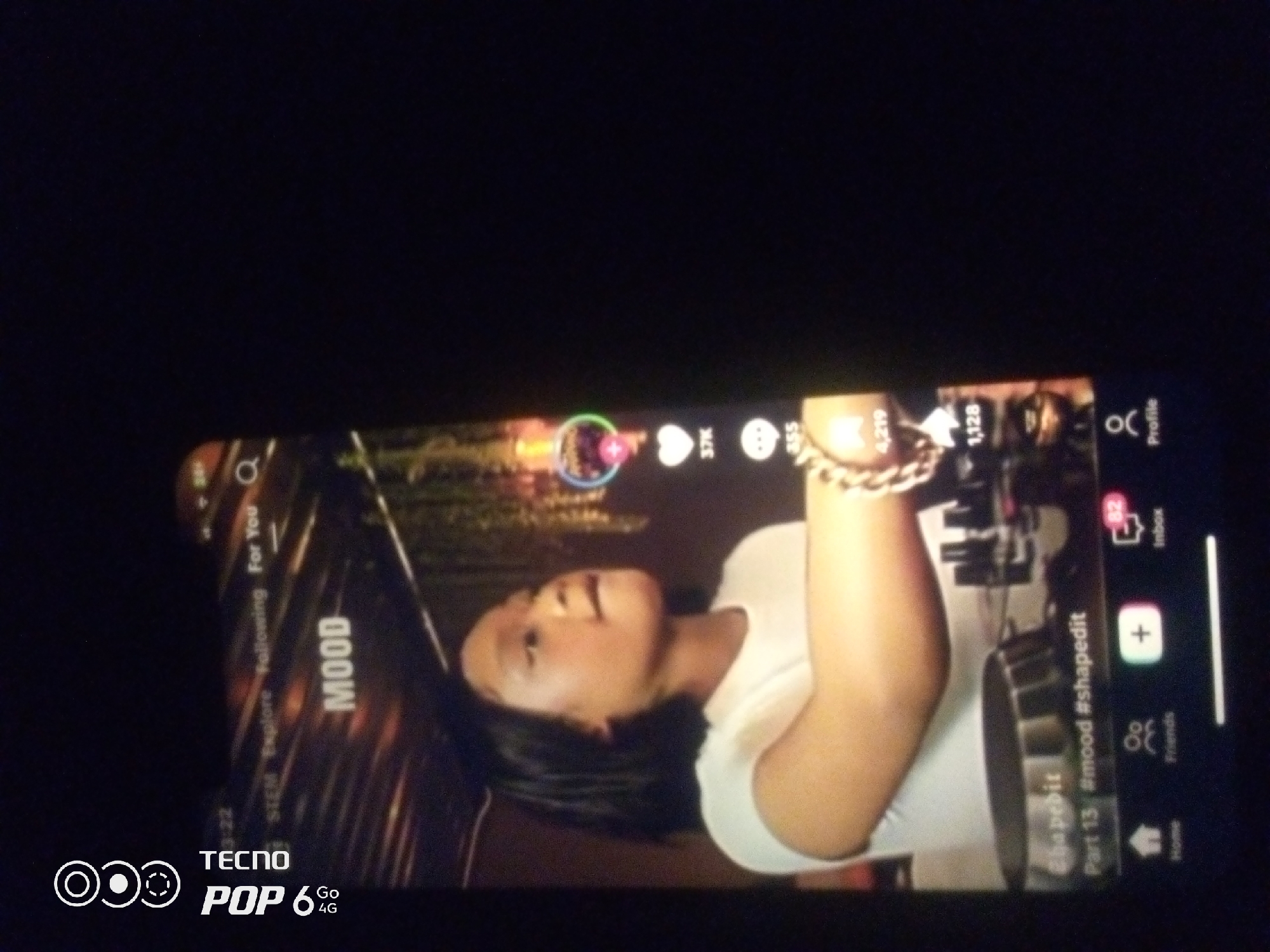 The image displays a close-up of a smartphone screen, oriented vertically and slightly tilted. The screen shows a video playing, featuring a young woman with dark hair and fair skin. She is wearing a white t-shirt and a gold chain necklace. The video appears to be from a social media platform, as indicated by icons for likes, comments, shares, and profile views, along with text overlays that include "MOOD," "@shapeedit," and "#mood #shapedit Part 13". The background of the video shows an outdoor or studio setting with blurred lights. The phone itself is black and appears to be a Tecno Pop 6 4G model, as a logo is visible at the bottom left of the screen. The overall scene suggests the use of a smartphone for entertainment or content creation in an indoor environment. There is no discernible information within the image that would indicate the location of Howayi, Nigeria.