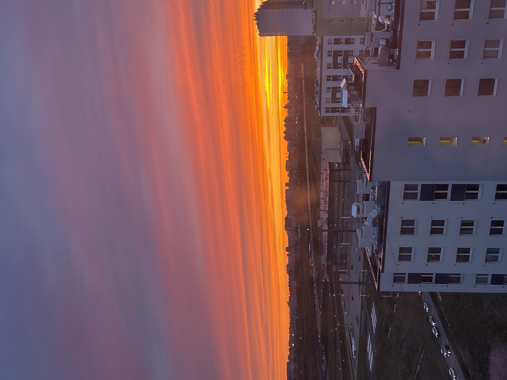 Captured from an elevated vantage point in Vsevolozhsk, Russia, the image presents a twilight urban panorama. The sky is ablaze with vibrant hues, transitioning from a fiery orange-red near the horizon to a soft purple at the top. This colorful sky is partially obscured by the distant cityscape.
Prominent in the right foreground are multi-story buildings, their facades marked with rows of windows. The buildings give way to streets with vehicle traffic and other urban elements like streetlights and pedestrian crossings. The setting appears to be at dusk as the light fades from the sky, creating a scene with a high-contrast, atmospheric blend of the natural and built environment.
