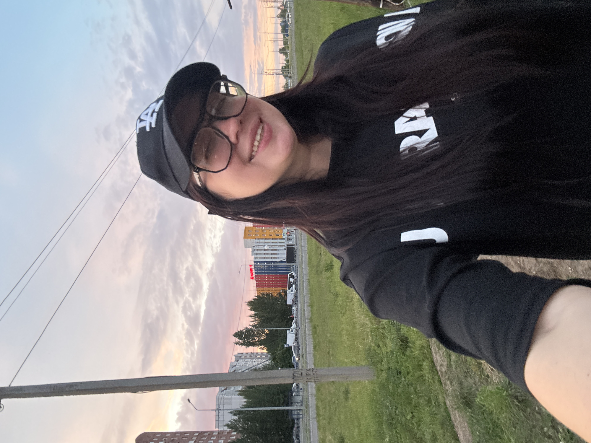 An image taken outdoors depicts a female subject in a selfie-style shot, smiling directly at the viewer. She has long, dark hair, is wearing a black baseball cap with a white "LA" logo, and large, clear-rimmed eyeglasses. Her upper body is visible, dressed in a black long-sleeved shirt with white text that partially reads "RAMONES" across the chest.
The background reveals an urban or suburban landscape at what appears to be sunset, characterized by a sky displaying hues of light blue, pink, and orange with scattered clouds. A prominent concrete utility pole stands vertically on the left side of the frame, showing faint alphanumeric markings. Behind the subject and to her left, a multistory residential building with reddish-brown brickwork is visible. Further back, a multi-lane road is populated with several cars, trucks, and a white bus, indicating traffic flow. Adjacent to the road, a strip of green grass and trees separates it from various buildings. One notable structure in the mid-ground features distinct vertical panels in red, blue, and yellow, and a red sign with white text, identifying it as a "MAGNIT" store, indicating the location is within the city of Unknown, Unknown. Further along the horizon, more buildings and additional utility poles with power lines are present, stretching into the distance under the fading daylight. The immediate foreground consists of green grass and a dirt path.