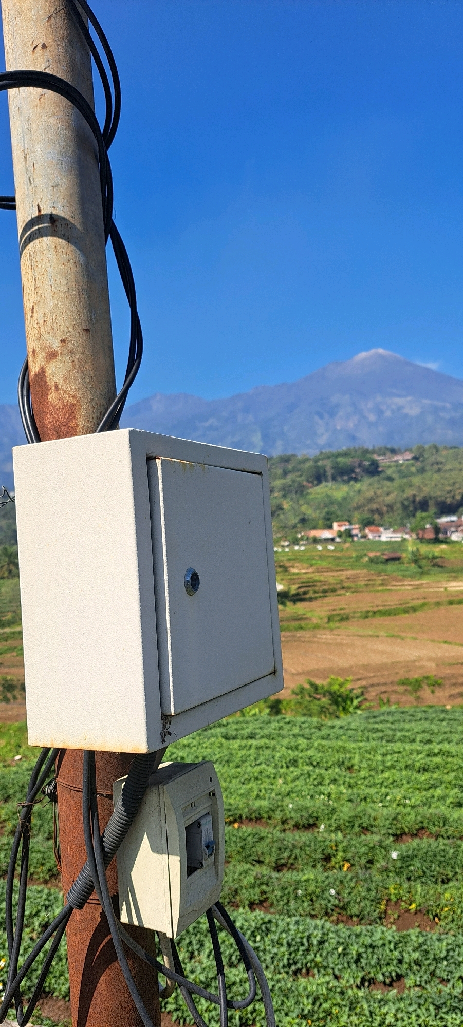 In Belik, Indonesia, a close-up, vertical shot captures a utility pole in the foreground. The pole, weathered and rusty, supports a white electrical box with a silver circular latch. Below this, a smaller, beige electrical enclosure is mounted, containing a circuit breaker. Black electrical cables snake around the pole and the enclosures. The background reveals a vibrant green agricultural field, likely cultivated with crops, stretching towards a village nestled among rolling hills. In the distance, a majestic mountain range dominates the horizon, its peaks veiled by a soft haze under a clear, bright blue sky. The scene is devoid of human presence, focusing solely on the infrastructure and natural landscape.