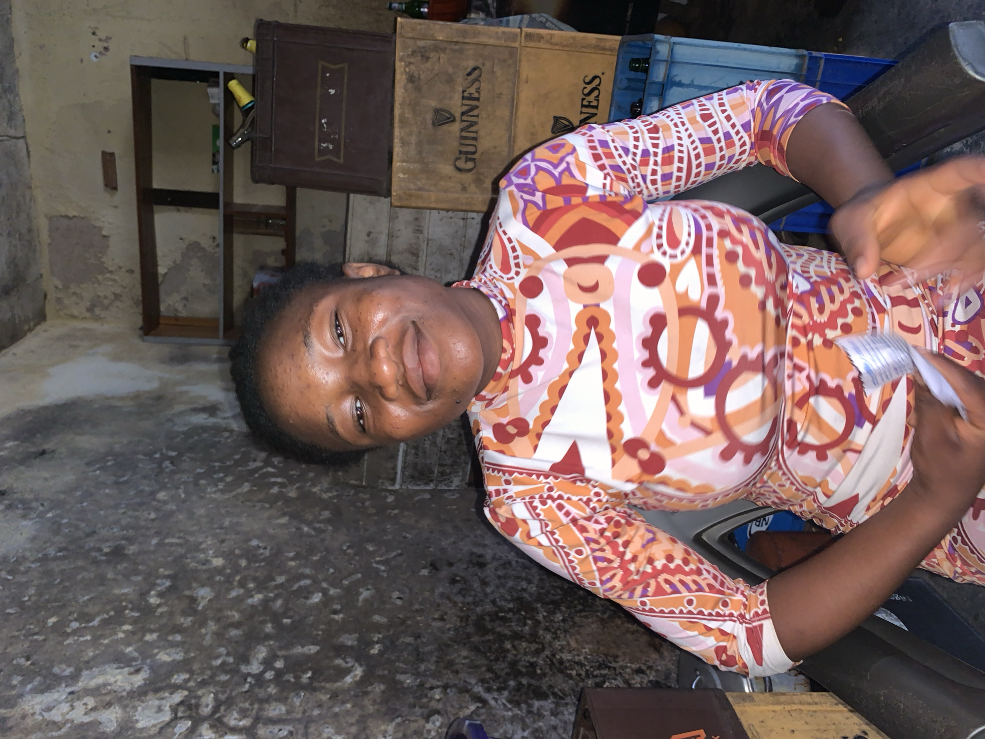 A medium shot depicts a dark-skinned woman with short, dark hair, facing forward and angled toward the camera. Her clothing is a patterned, long-sleeved garment in shades of orange, white, and purple. She holds a white object, possibly a wrapper. The environment includes a dark, textured floor and walls that appear aged and stained. Behind the woman are stacked wooden crates labeled "GUINNESS" and a blue plastic crate with bottles. A dark wooden frame is visible on the left side. The location may be Jos, Nigeria, based on the prompt.