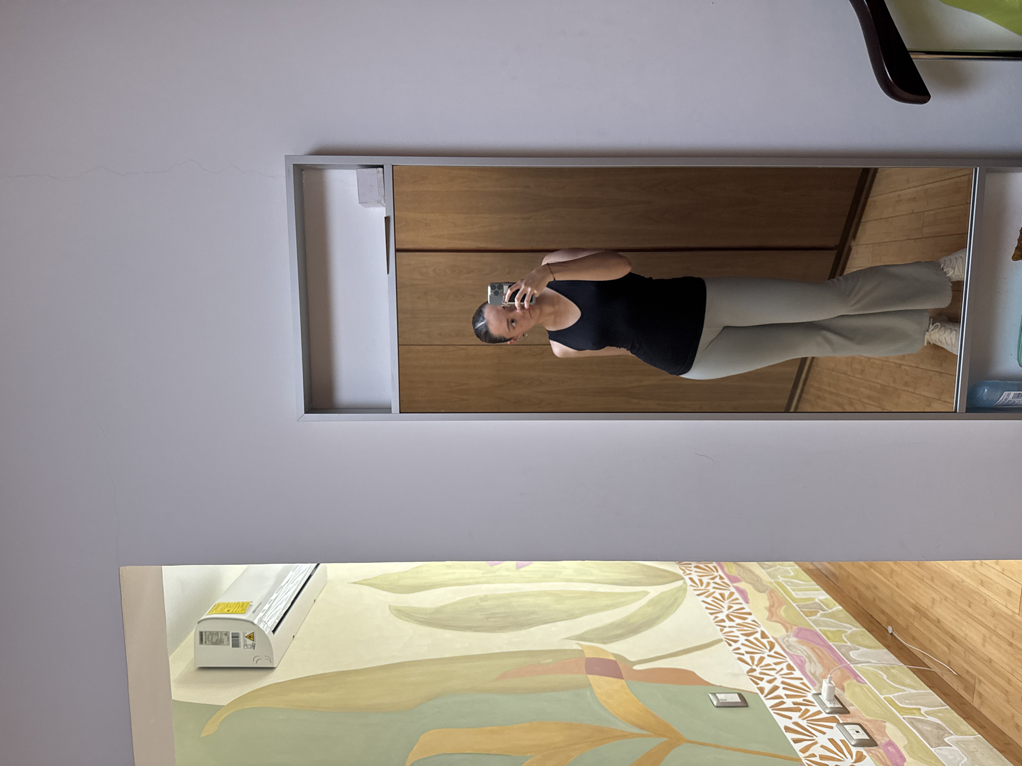 The image displays an indoor scene within San Bernardino Tlaxcalancingo, Mexico, centered around a tall, framed mirror on a light grey wall. Reflected in the mirror is a single individual, appearing female, dressed in a black V-neck tank top, light grey flared pants, and light-colored athletic shoes. She holds a light-colored smartphone in her raised right hand, positioned to capture a mirror selfie, with the phone partially obscuring her face. Her hair is pulled back, and small earrings are visible.
The reflection behind the individual shows a light brown wooden wardrobe or closet with two doors. Above the reflected head, a small white rectangular object rests on a shelf integrated into the mirror's frame. Below the reflection, a blue bottle and a green object are partially visible on a lower shelf.
To the immediate left of the mirror, a wall is decorated with a large, abstract botanical mural in tones of light green, pale yellow, and terracotta. Below this mural, an orange and white patterned border runs horizontally, followed by a section featuring pink, light green, and light brown geometric patterns. A white light switch is mounted on the mural, and two white electrical outlets are visible lower on the wall, with a white charging cable plugged into the lower outlet and extending downwards.
High on the left wall, a white split-system air conditioning unit is mounted, displaying a yellow warning label and other operational labels. The ceiling is light grey and exhibits a thin, vertical crack above the mirror's top edge. The flooring throughout the visible area, including its reflection, consists of light brown hardwood planks. In the lower right corner, outside the mirror's frame, a dark brown, curved armrest of a chair or similar furniture item is partially visible.