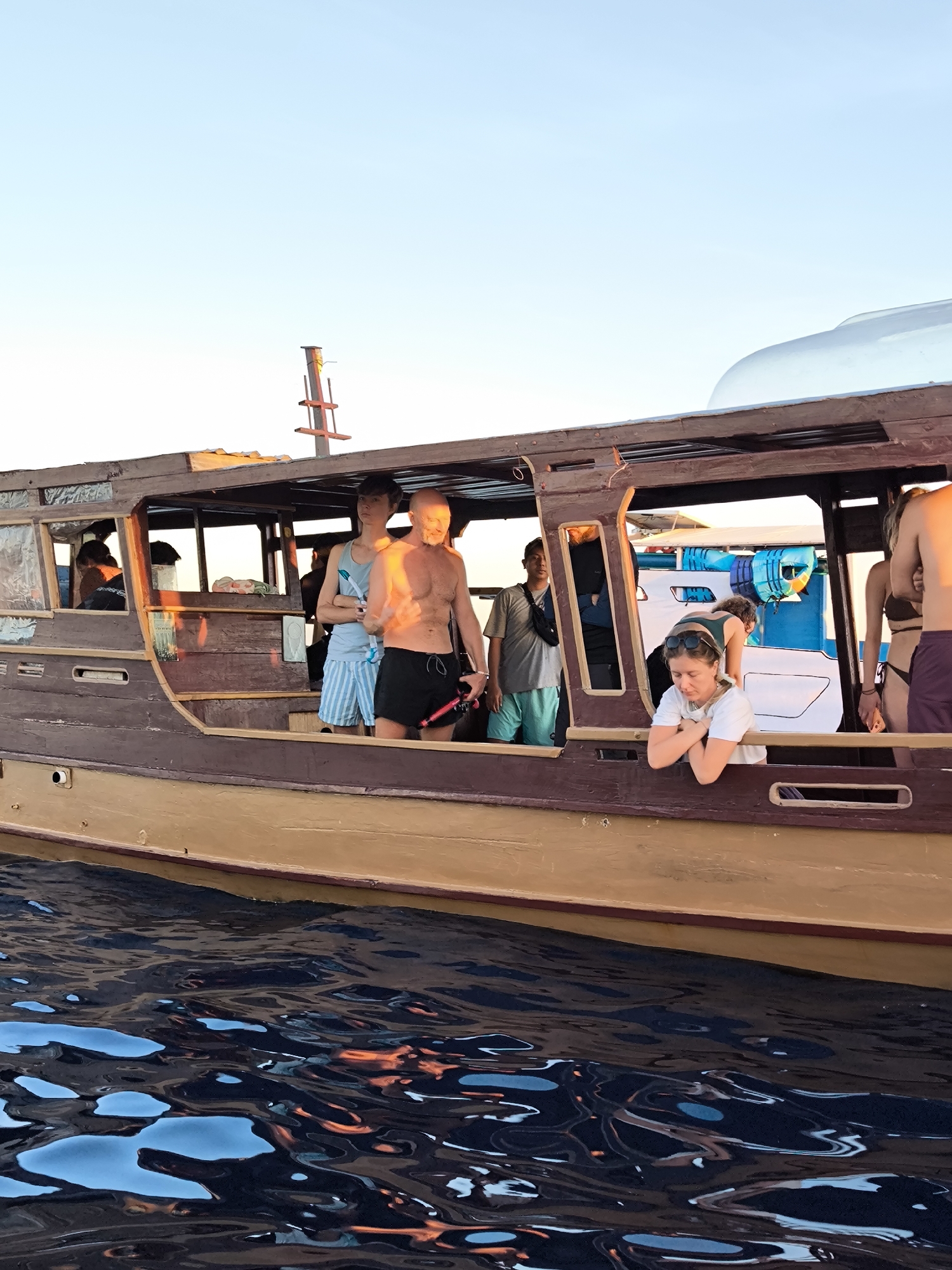 On a boat in Unknown, Indonesia, several individuals are present. An older man with a bare chest and dark shorts holds a snorkeling mask. A woman in a white shirt leans out of a window with her hands clasped under her chin. Other individuals can be seen inside the boat, some in swimwear. The boat has a brown wooden exterior and is on water with reflected sunlight.